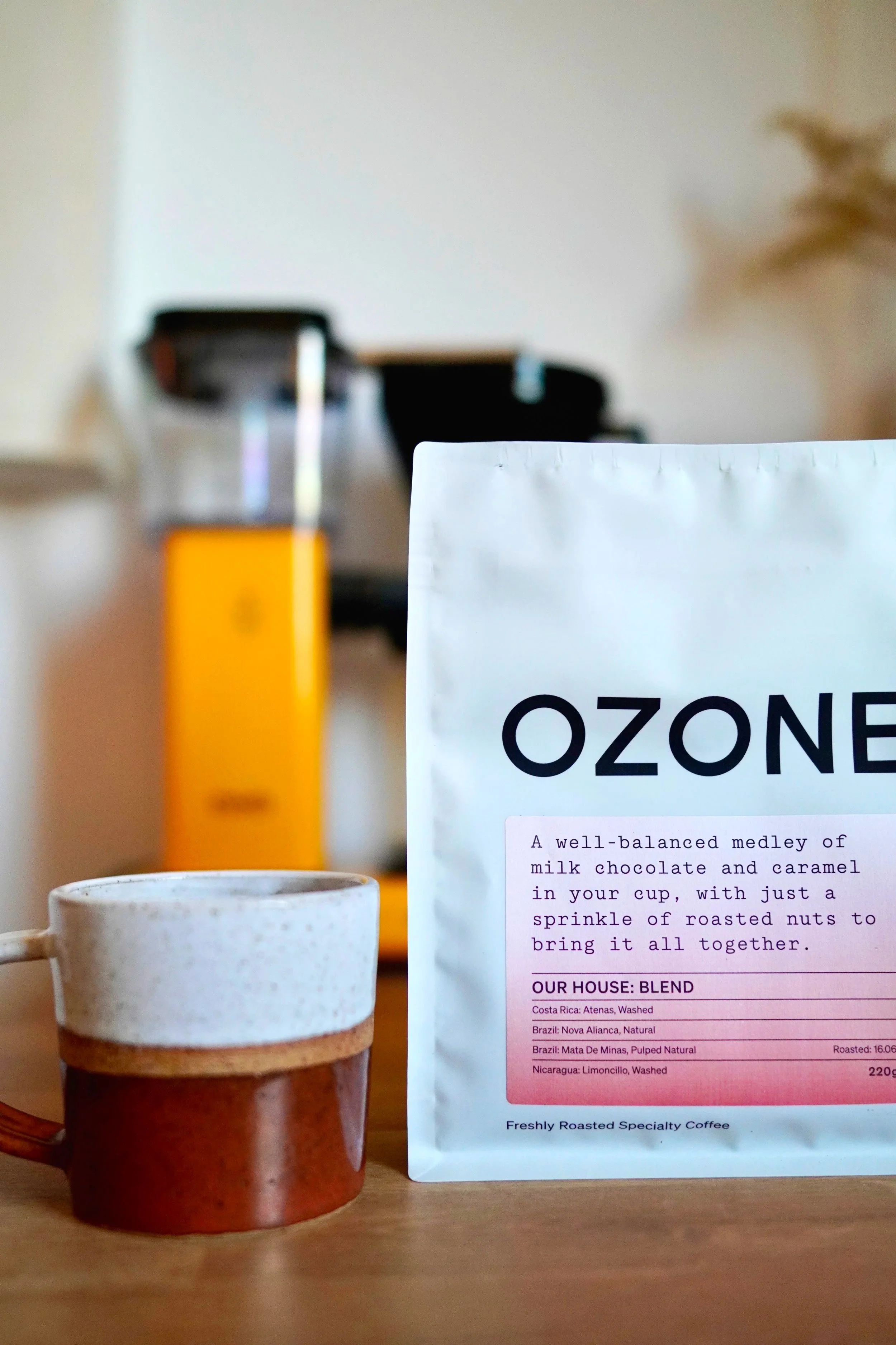 A white bag of coffee labeled 'OZONE' with a description of a coffee blend, positioned in front of a layered ceramic mug and a yellow machine in the background.