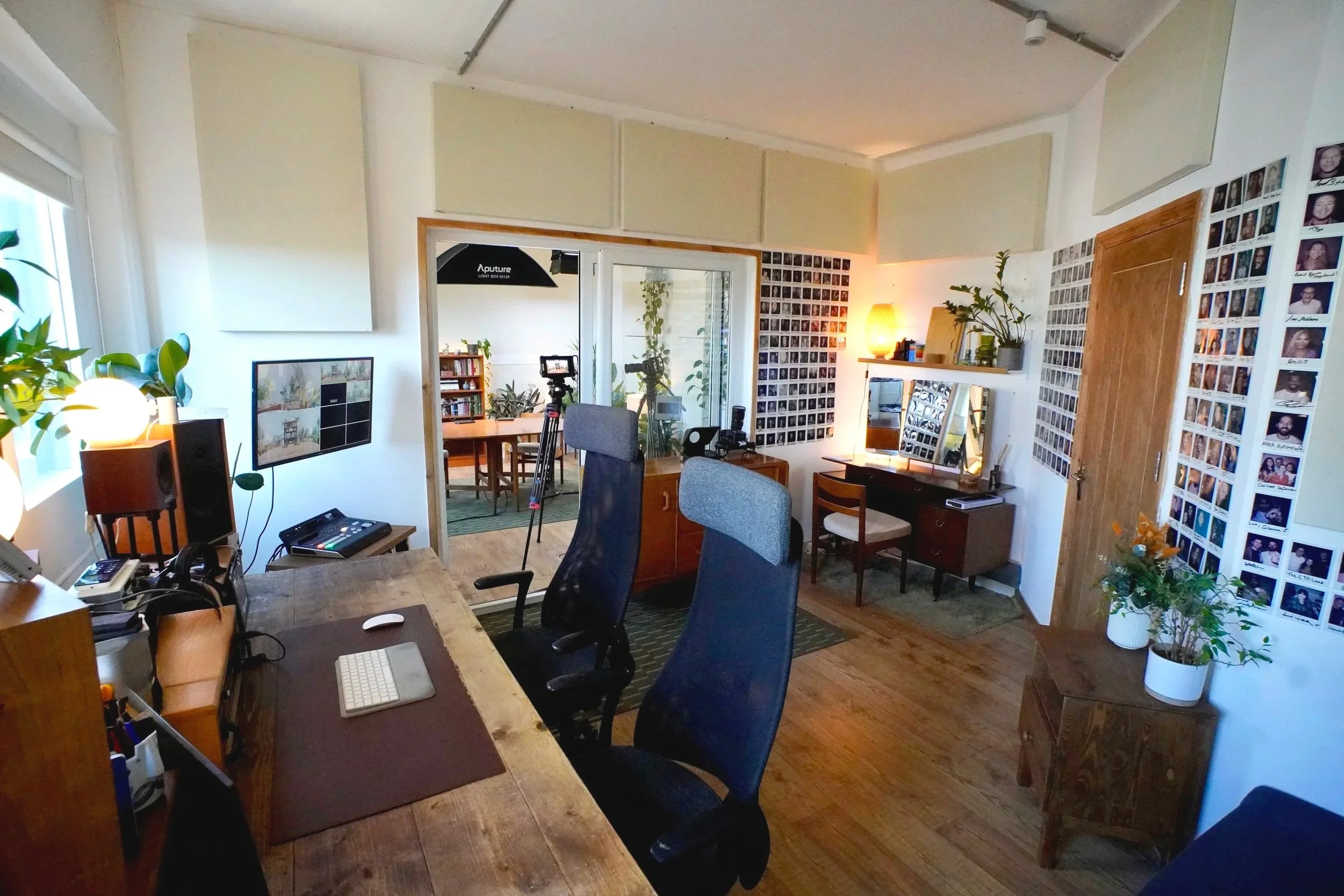 A cozy home office with a wooden desk, multiple chairs, and wall decorations including photo collages and plants, illuminated by warm lighting.