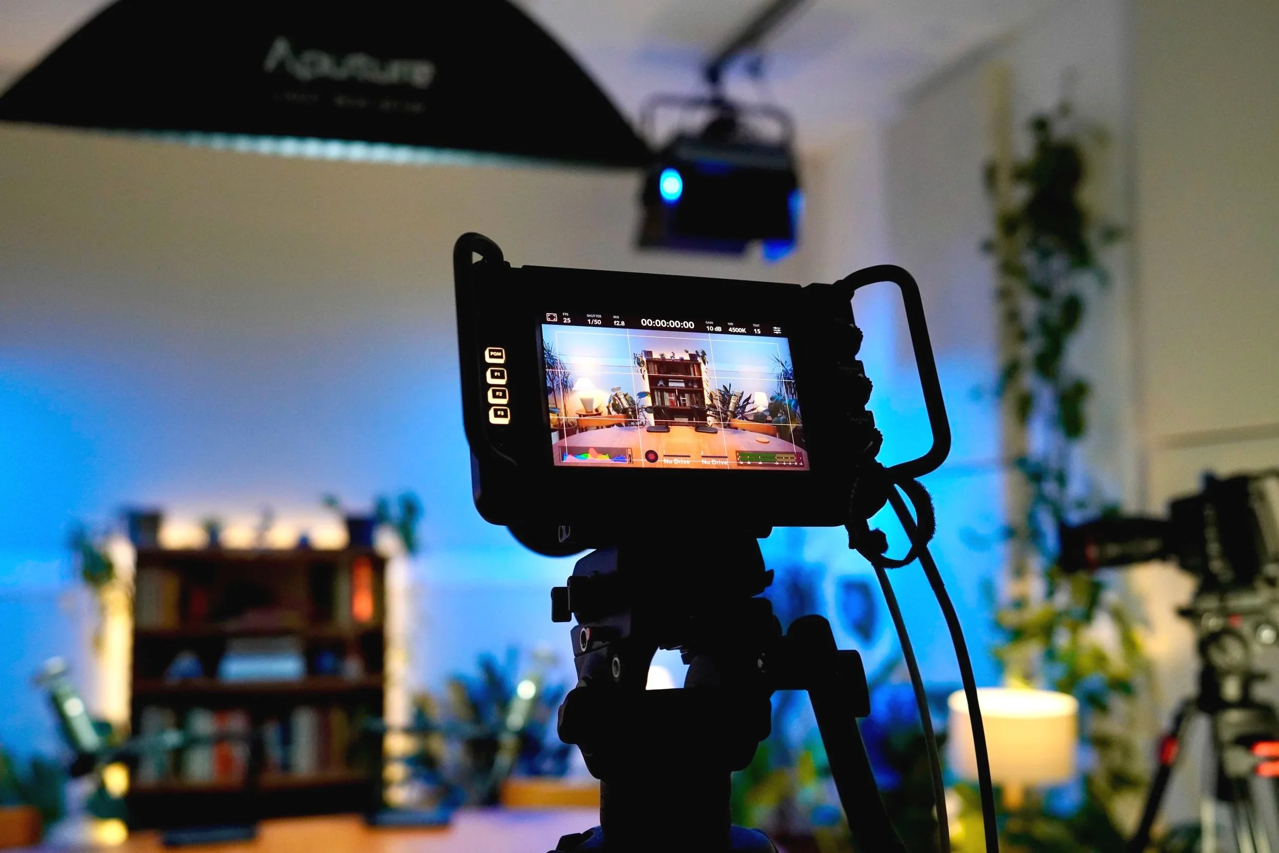 Close-up of a digital camera on a tripod capturing a scene in a room with a bookshelf and plants, illuminated by blue lighting.