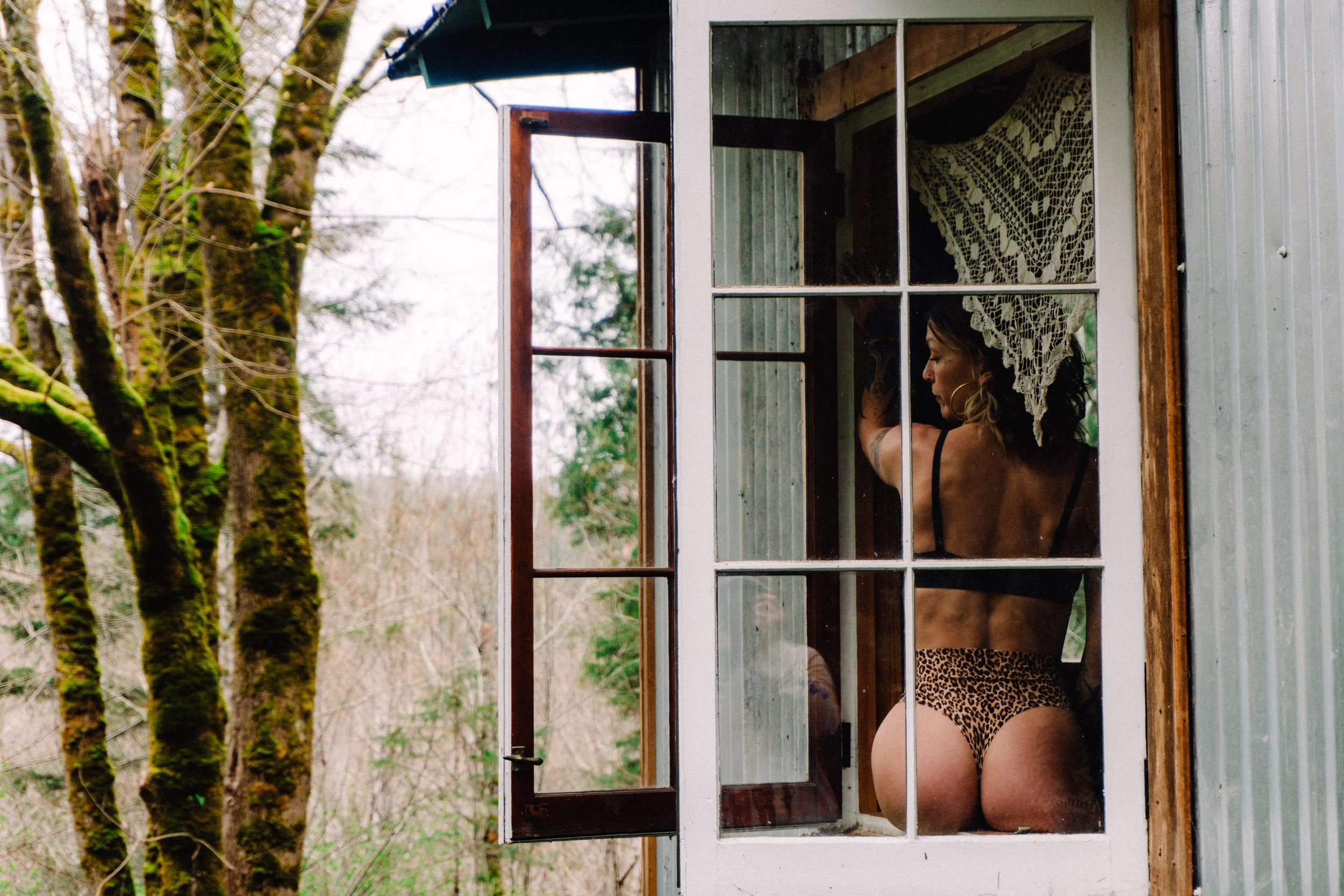 A woman with dark hair and wearing leopard print underwear, seen through the window of a small wooden and metal shed, outside in a wooded area.
