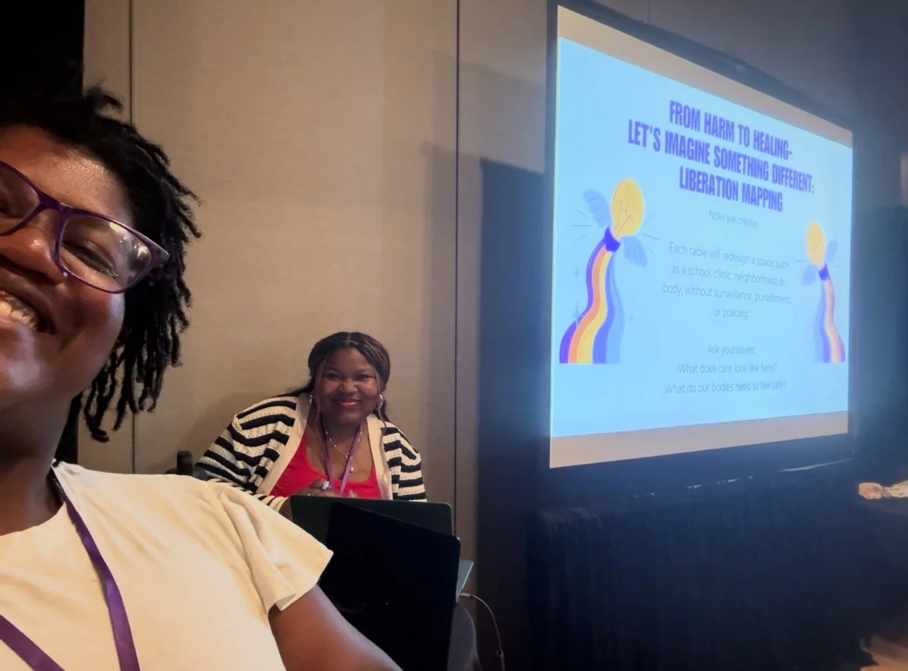 Two women smiling at a conference or workshop, with a presentation slide projected on the wall behind them. The slide discusses transitioning from harm to healing and features illustrations of light bulbs with rainbow-colored streams.