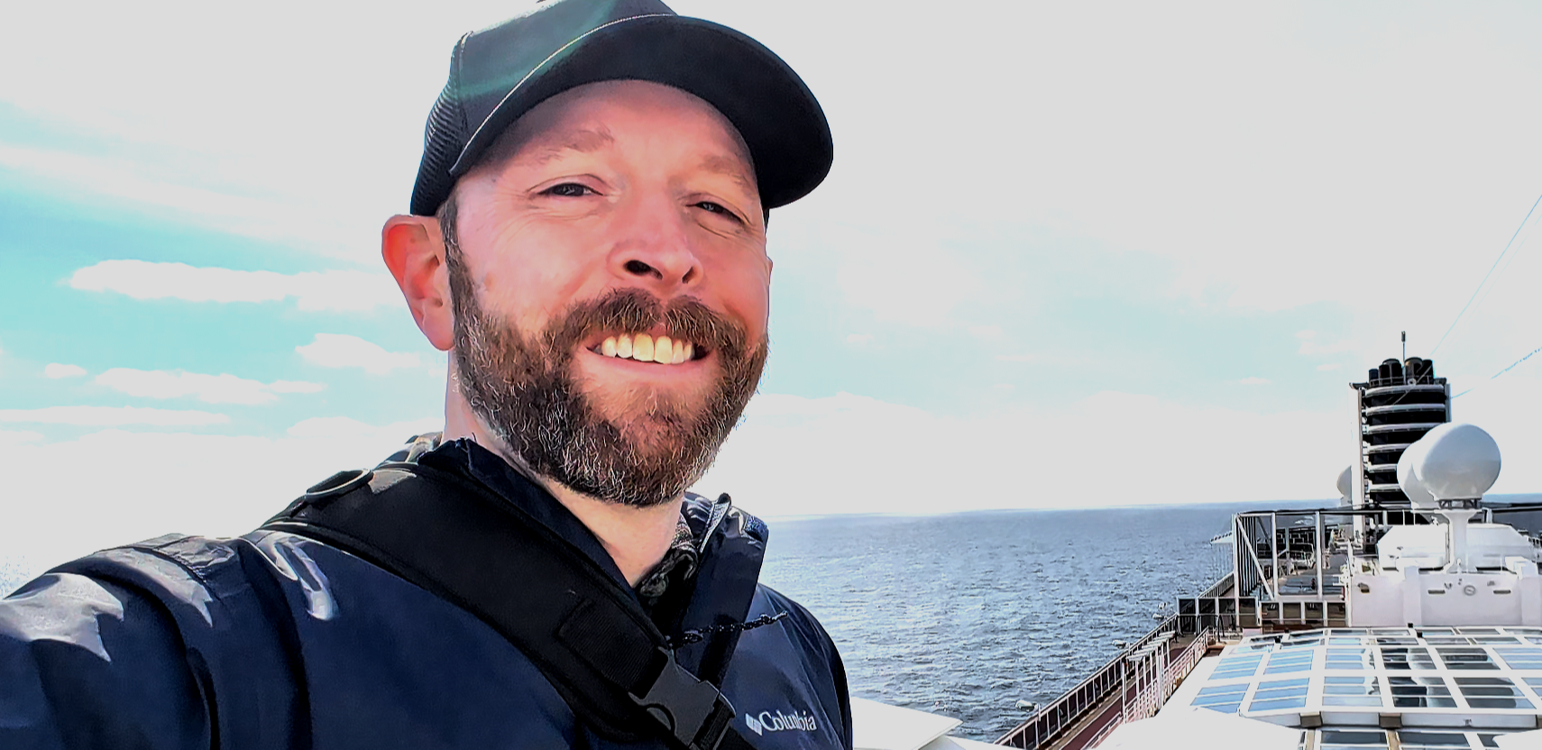 Man with beard smiling on ship deck, ocean and sky in background.