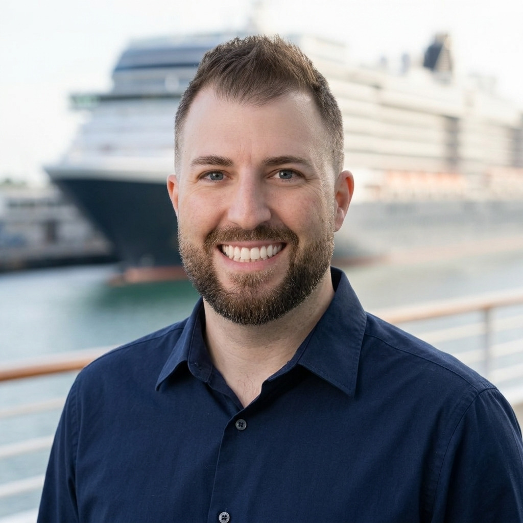 Justin, independent travel agent. A smiling man with short brown hair and a beard, wearing a navy blue shirt, standing outdoors with a large cruise ship in the background.