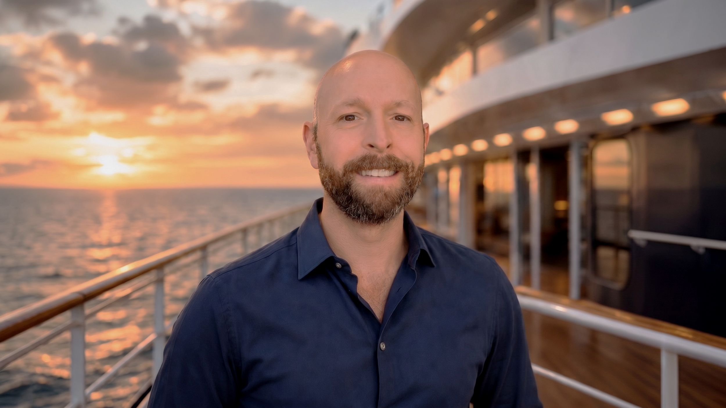 Brad, independent travel agent of Brad On Board. A smiling man with a beard and a dark blue shirt standing on the deck of a cruise ship at sunset, with the ocean and cloudy sky in the background.