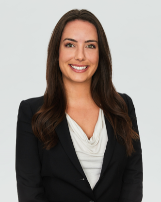 A young woman with long brown hair, smiling, wearing a black blazer over a white top against a plain light background.