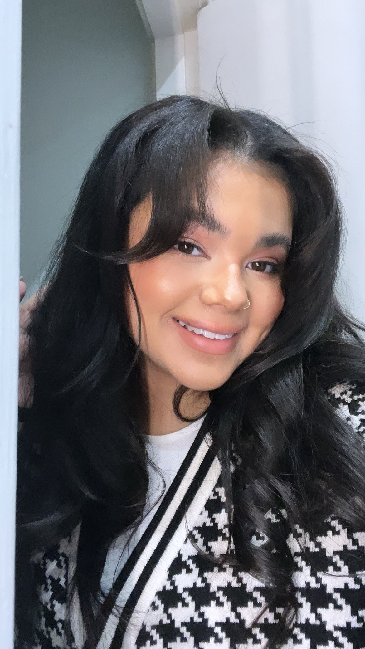 A young woman with long black wavy hair, smiling, wearing a black and white patterned jacket, with a nose ring, in front of a mirror.