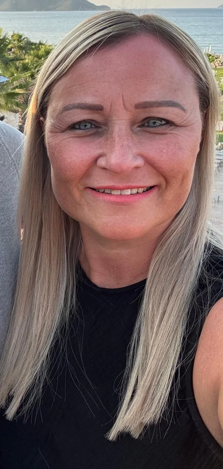 Close-up of a middle-aged woman with blonde hair and blue eyes smiling outdoors near the water with greenery in the background.