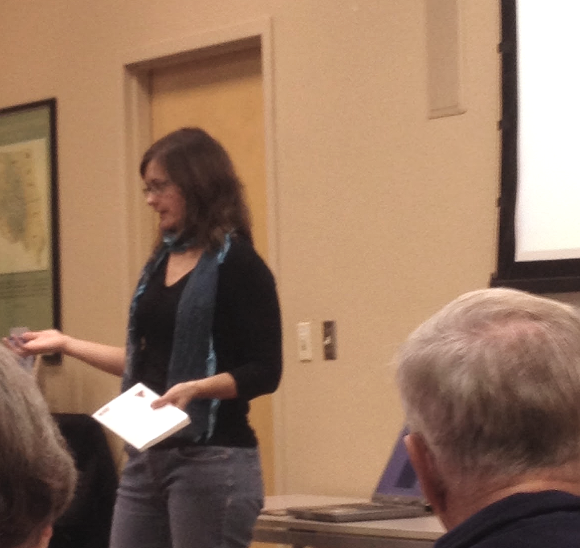 A woman giving a presentation in a conference room, holding a phone and paper, with attendees visible in the foreground.
