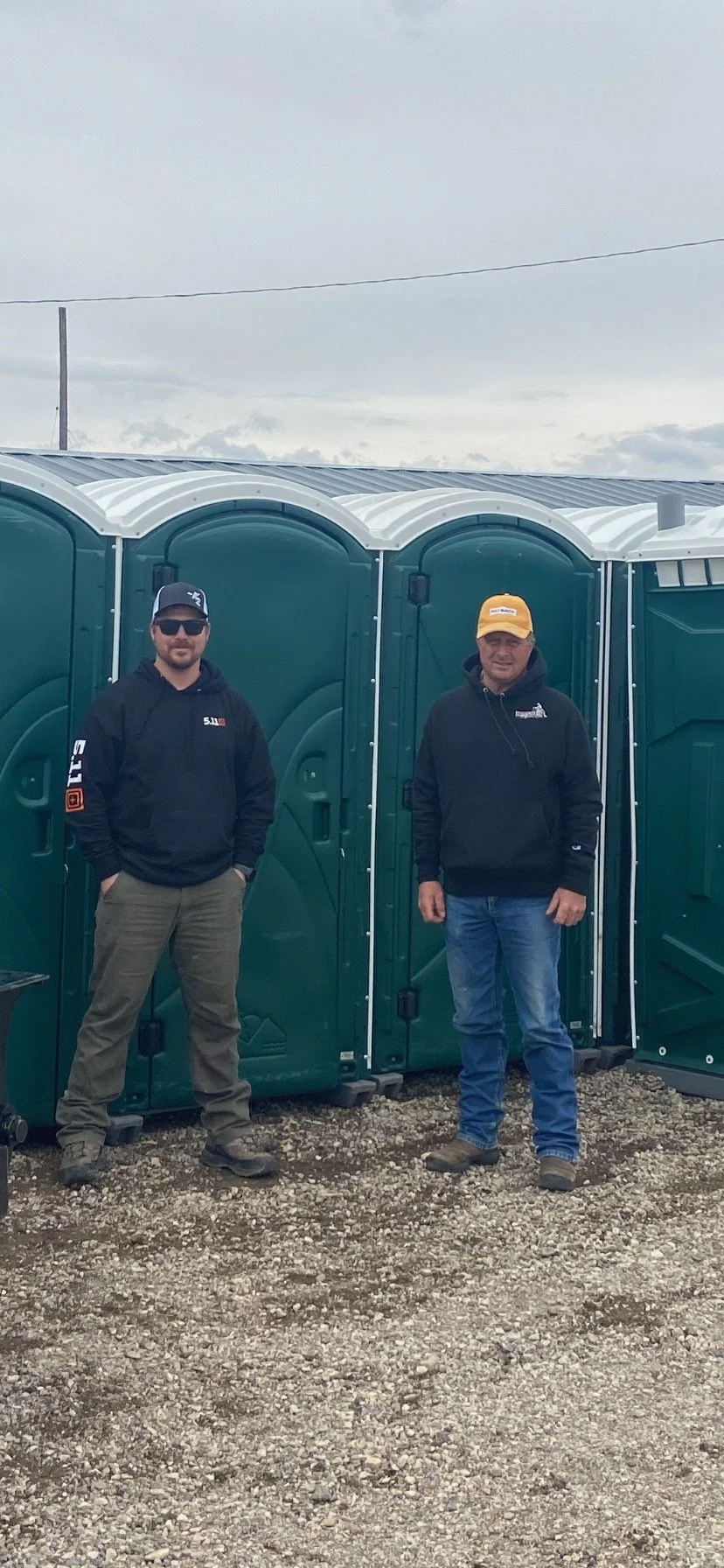 Two men standing outside in front of green portable toilets, wearing hoodies and hats.