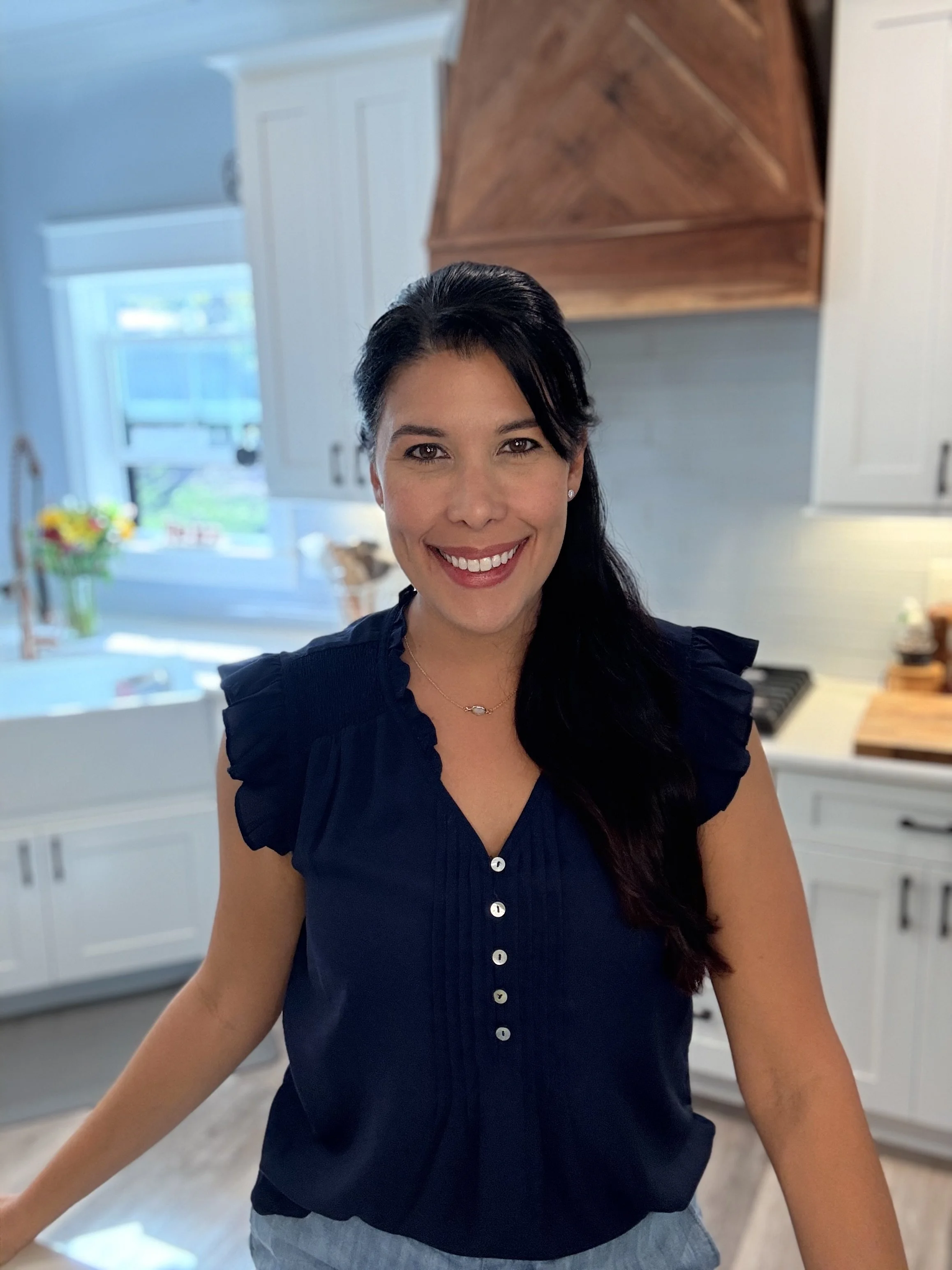 A woman with long dark hair, wearing a navy blue blouse with ruffled sleeves and buttons, smiling in a bright, white kitchen with natural light, a window, and kitchen cabinets in the background.