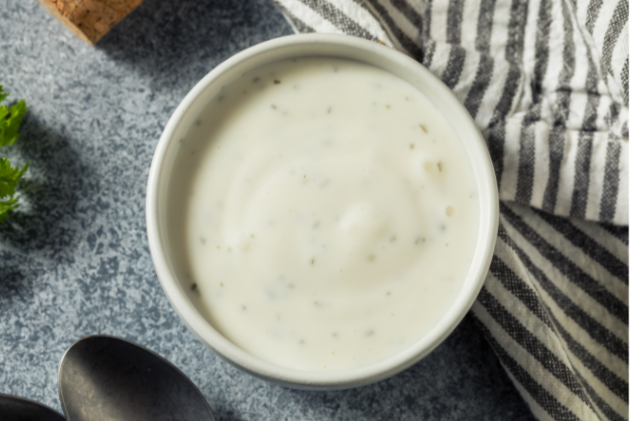 Close-up of a bowl of creamy ranch dressing with black pepper pieces, on a gray textured surface with a striped napkin nearby.