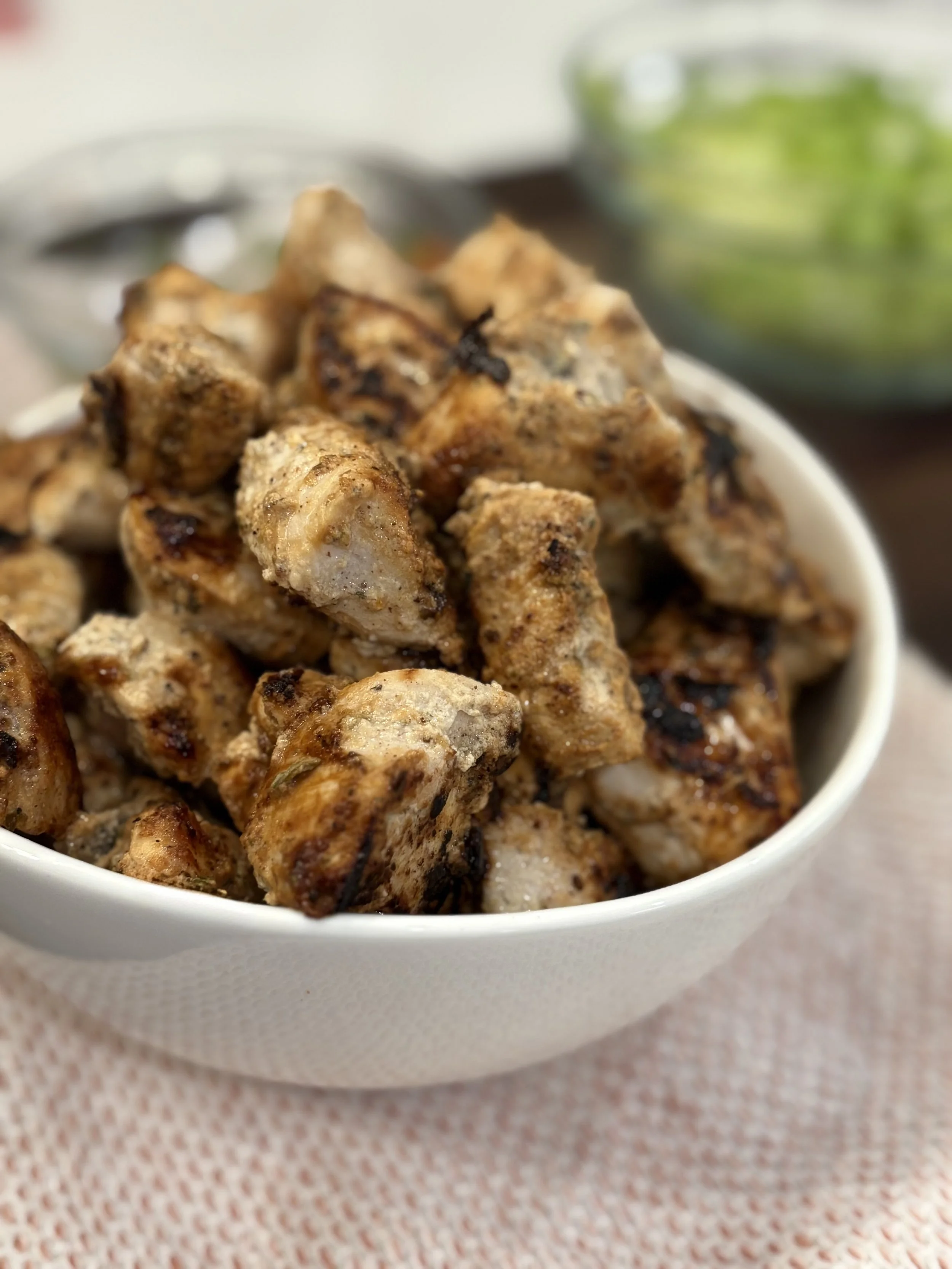Close-up of a white bowl filled with grilled chicken chunks, garnished with spices, placed on a pinkish cloth. In the background, there is a blurred bowl of salad.