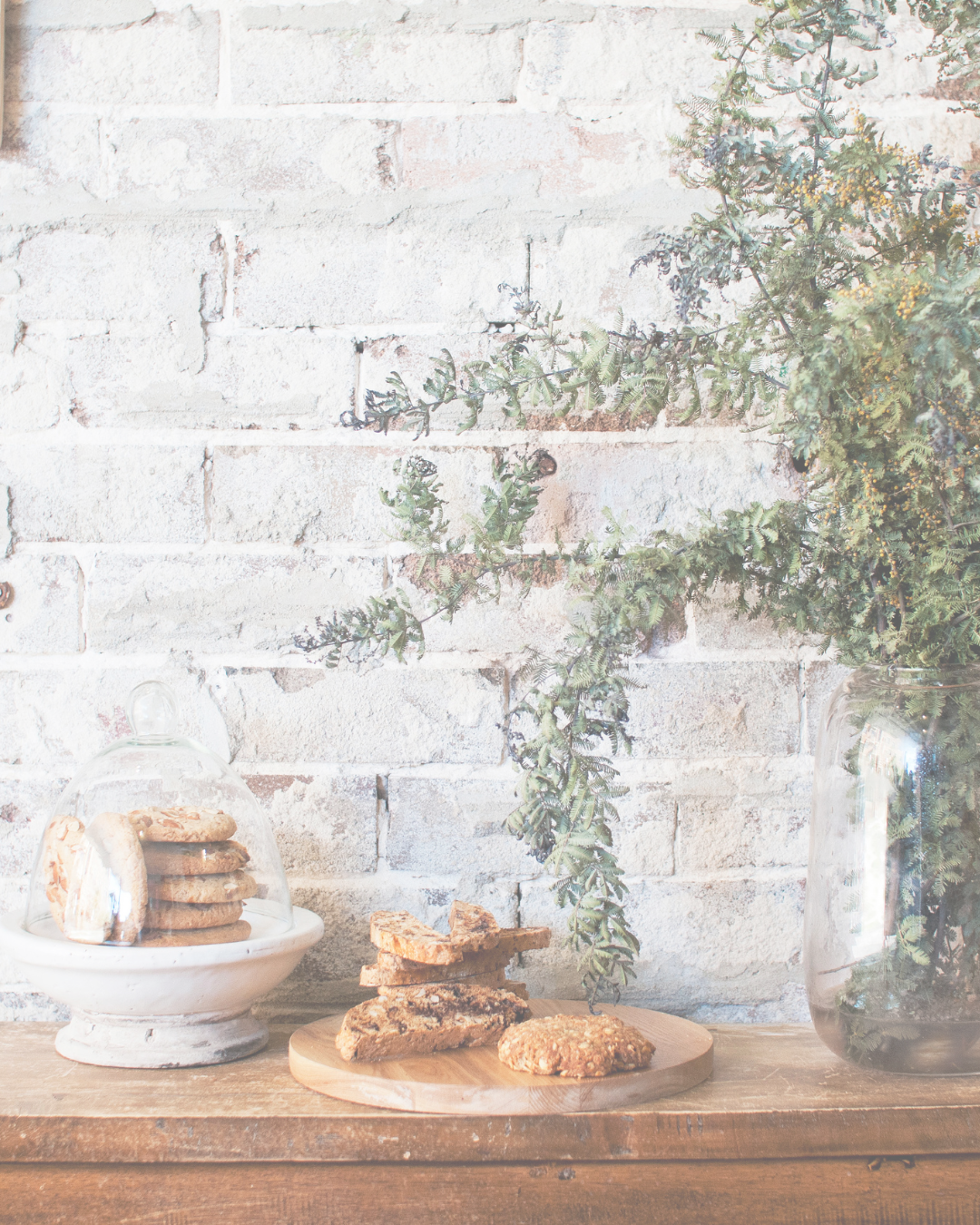 Cookies on a white cake stand with a glass dome, on a wooden table next to a small pile of baked goods on a round wooden board, and a large glass jar filled with green foliage against a white brick wall.