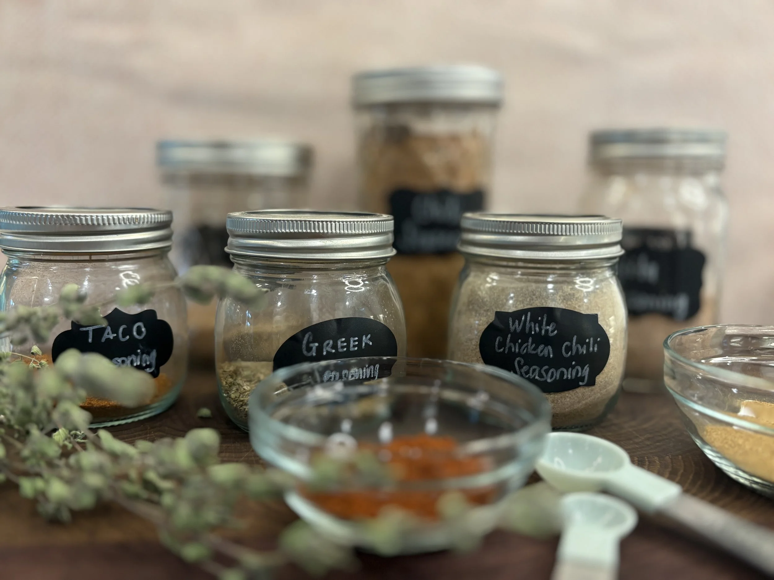 Several glass jars with metal lids labeled with spice names, including taco seasoning, Greek seasoning, and white chicken chili seasoning, on a wooden surface with measuring spoons and spice bowls.