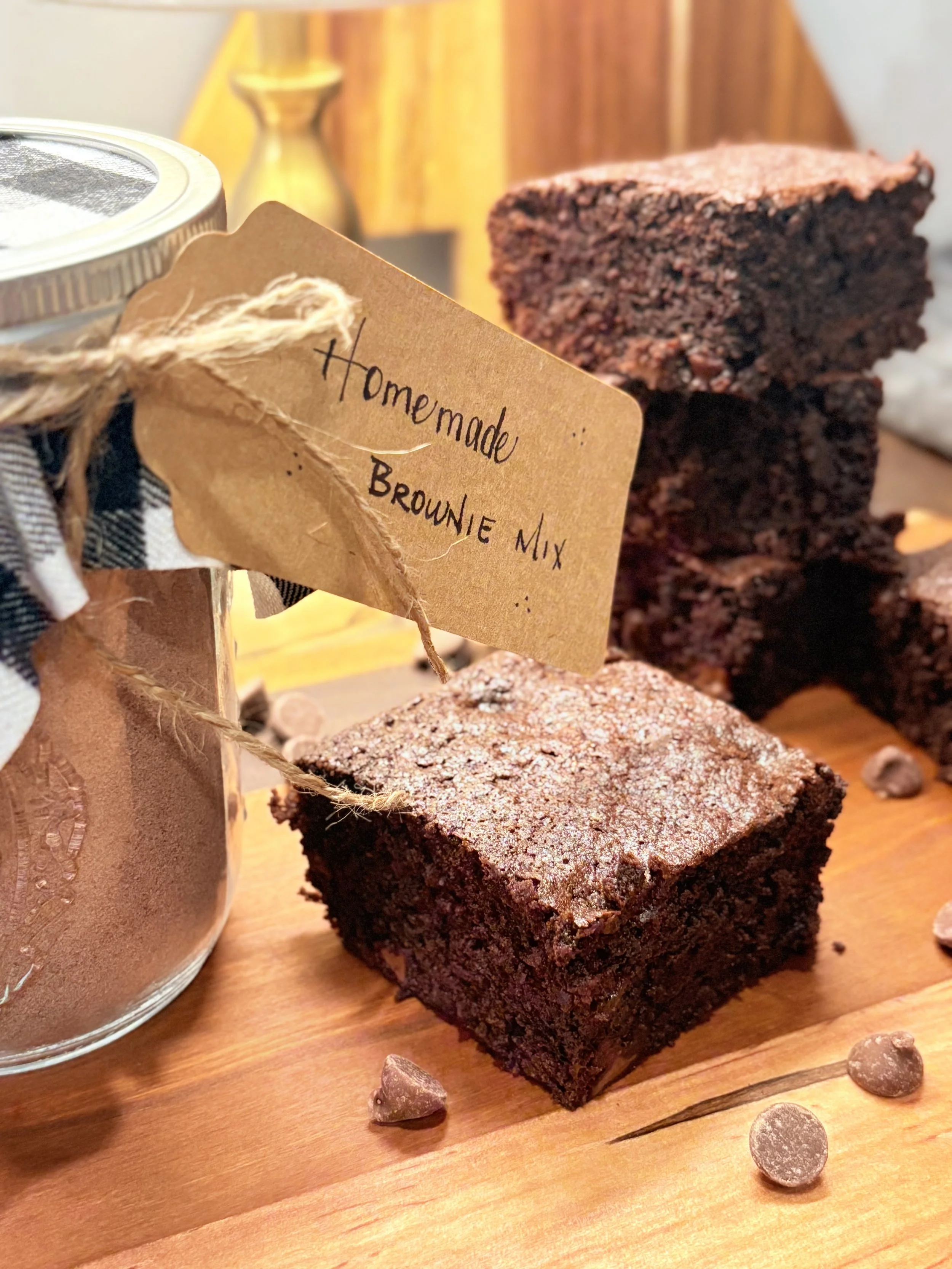 A close-up of a homemade brownie square with a rough top crust, surrounded by additional brownie pieces and chocolate chips on a wooden surface. A jar with a checkered cloth and a handwritten tag reading 'Homemade Brownie Mix' is also in the scene, with a blurred background.