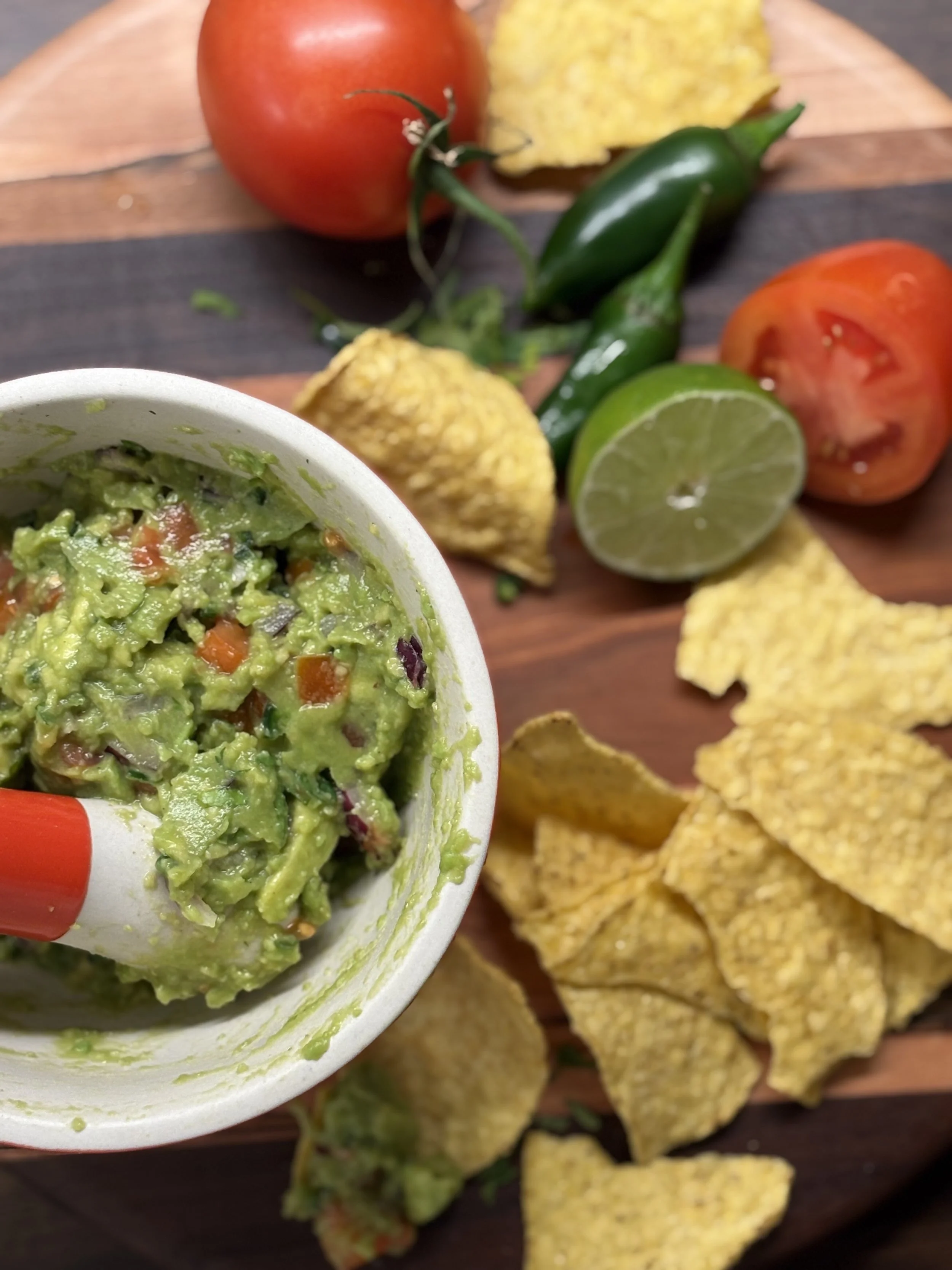 Close-up of a cup of guacamole with a chip, surrounded by fresh tomatoes, jalapeño peppers, lime, and corn chips on a wooden surface.