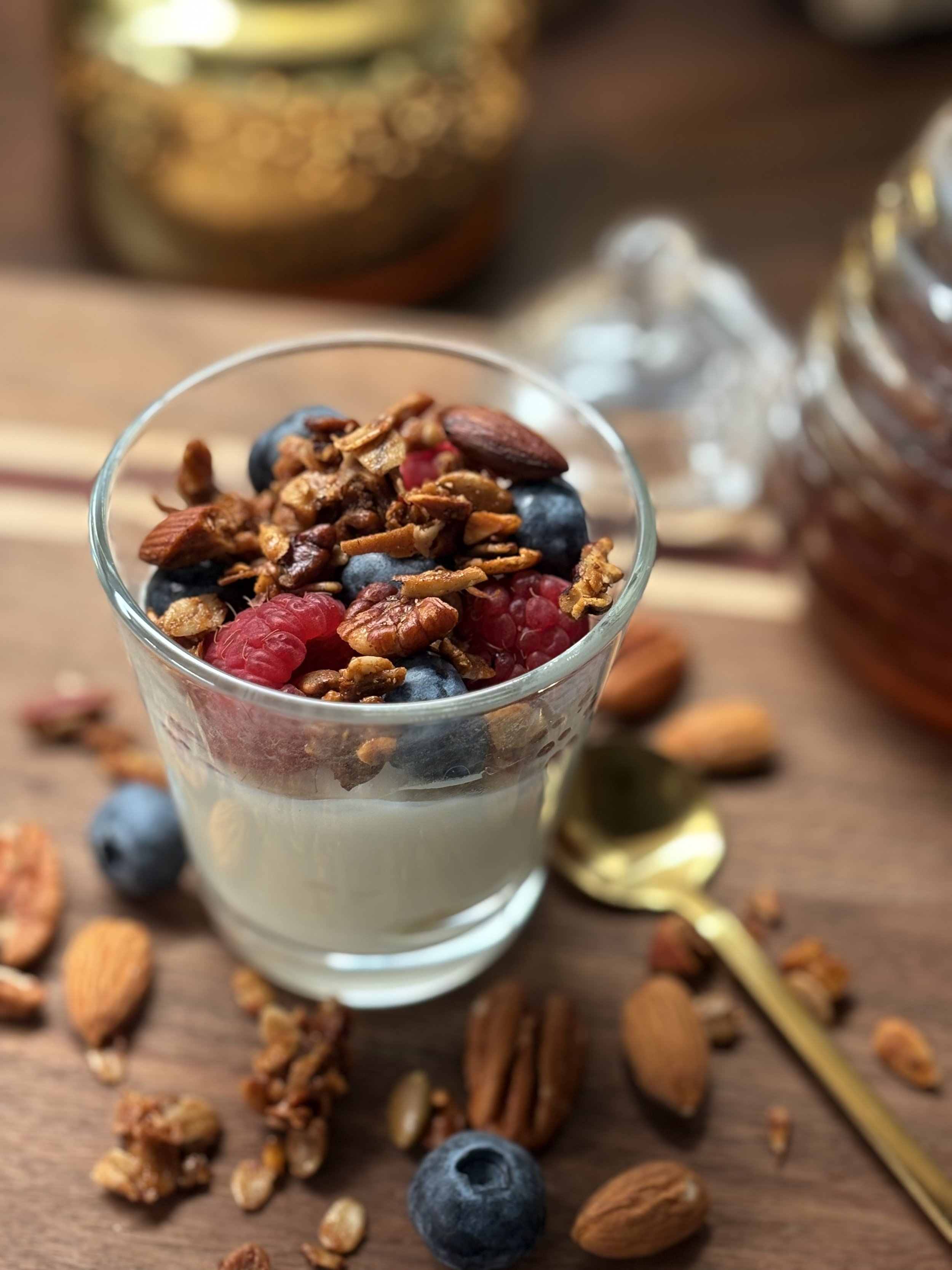 Yogurt parfait topped with mixed berries and granola, with scattered almonds and blueberries on a wooden surface.
