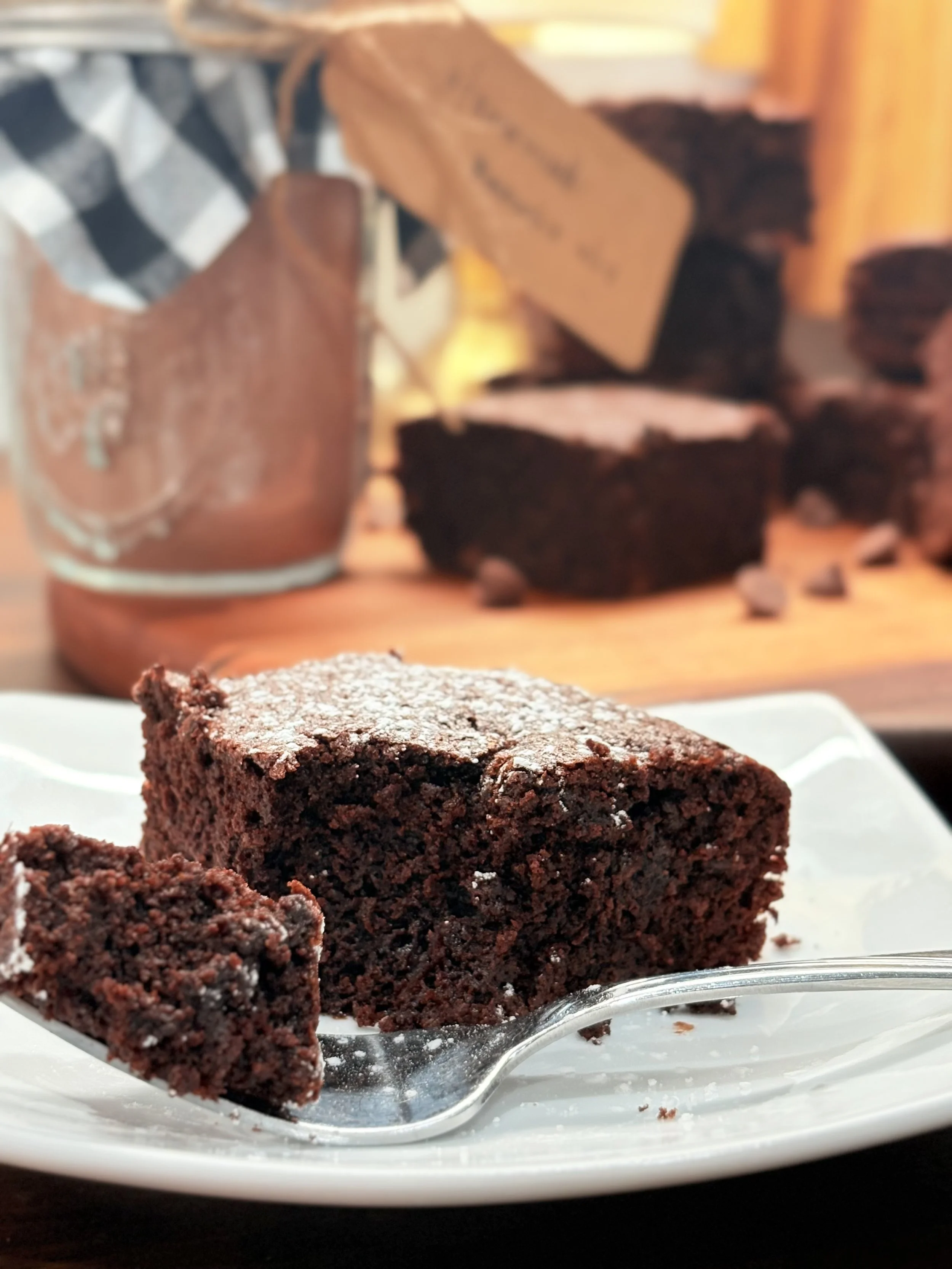 A slice of chocolate cake with powdered sugar on top served on a white plate, with a fork beside it, and a blurred background of a chocolate cake and a jug.