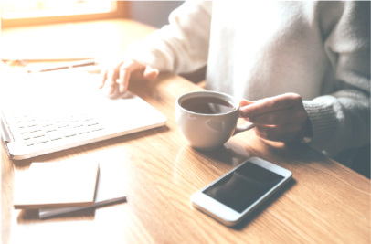 Person with gray sweater sitting at wooden table with laptop, smartphone, notepad, and holding a coffee mug.