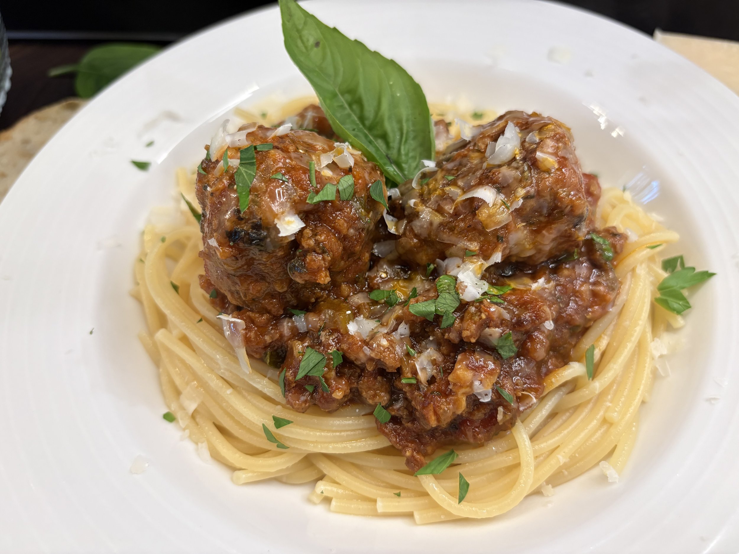 Spaghetti with meatballs topped with tomato sauce, chopped herbs, and grated cheese, garnished with a basil leaf on a white plate.