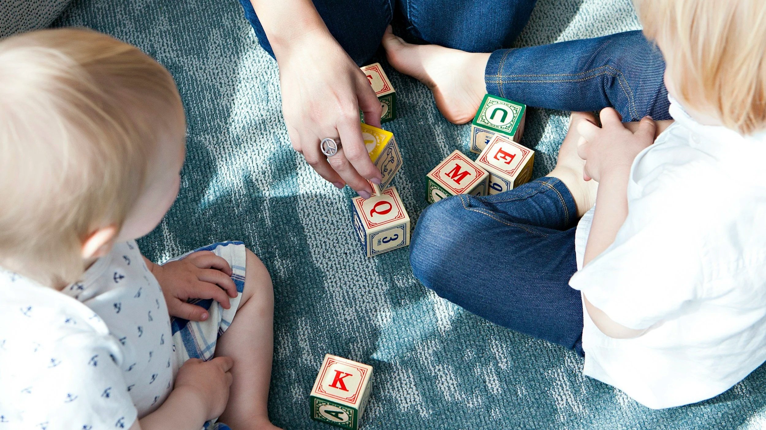 Two children sitting on a blue carpet playing with colorful alphabet blocks that spell out words. An adult is helping arrange the blocks.