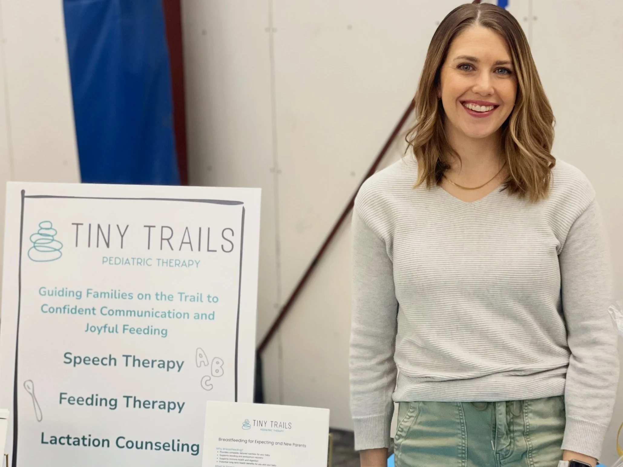 A smiling woman with shoulder-length brown hair wearing a striped long-sleeve shirt standing next to a poster for Tiny Trails Pediatric Therapy, which offers guidance for families on communication and feeding for children.