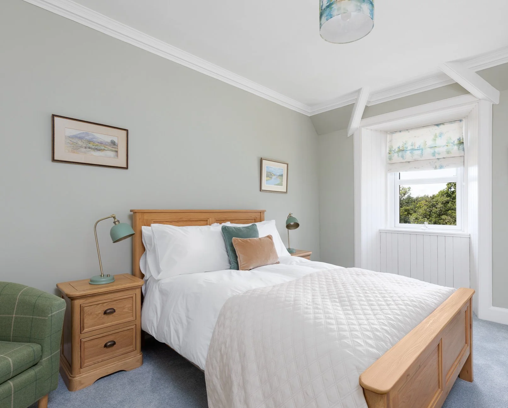 A cozy bedroom with a wooden bed frame, white bedding, and pillows. Two small wooden nightstands with green table lamps. A green plaid armchair with a brown pillow. Light gray walls, framed landscape art, and a window with a patterned roman shade, le