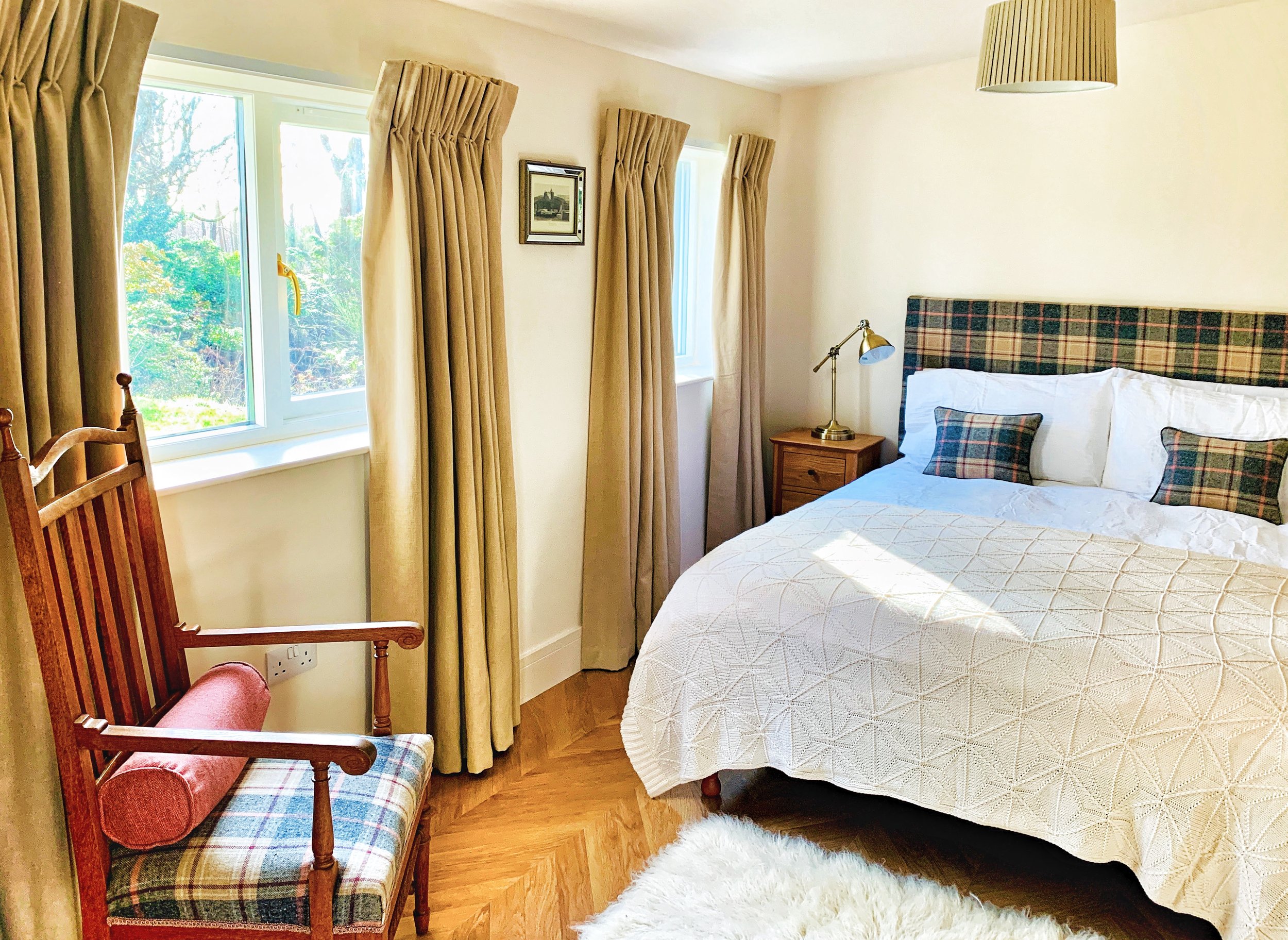 Living room with a light gray sofa, plaid cushions, a framed landscape photograph on a light green wall, a white side table with a lamp, a tripod floor lamp, and a plaid ottoman with a wooden tray holding cups and a magazine, all on a cream rug and hardwood floor.