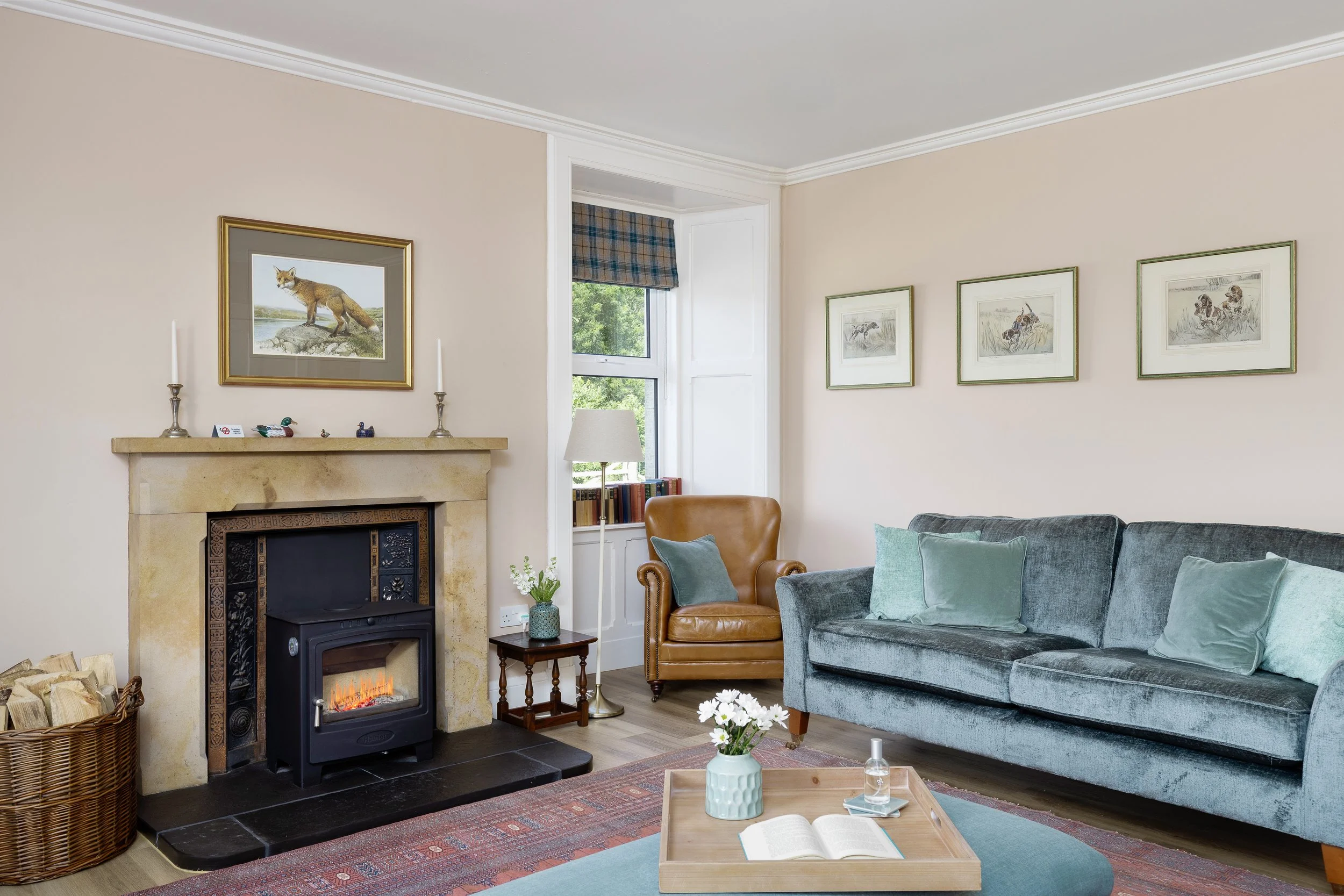 Living room with beige walls, large window with plaid shade, fireplace with a TV on a stand, blue upholstered couch and armchairs, a red patterned rug, and decorative pillows.