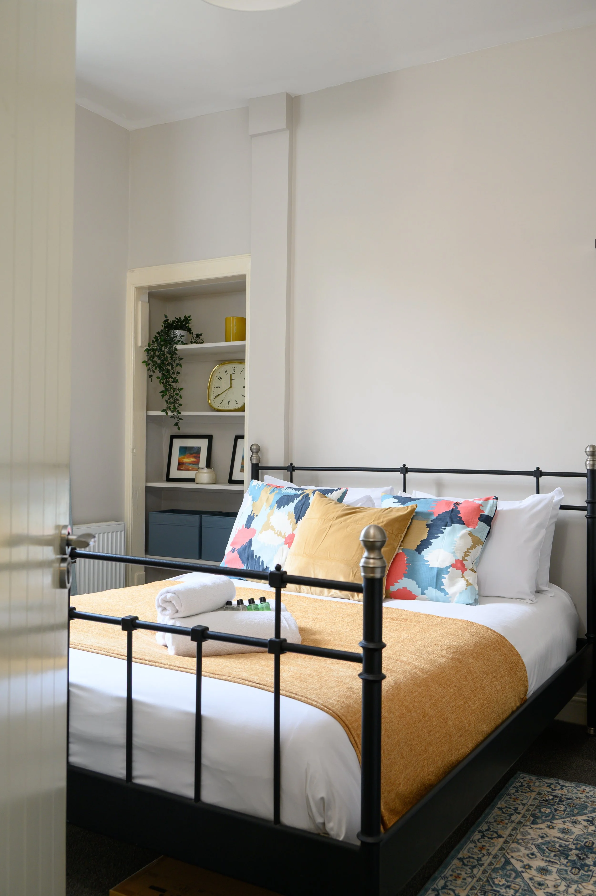 A cozy bedroom with a bed covered in white linens, pink and black pillows, and a pink throw blanket. There is a bedside black lamp on a small black table, and a desk with an orange chair and a book near the window. The room has pink curtains, a window revealing a brick building outside, and decorative framed pictures on the wall.
