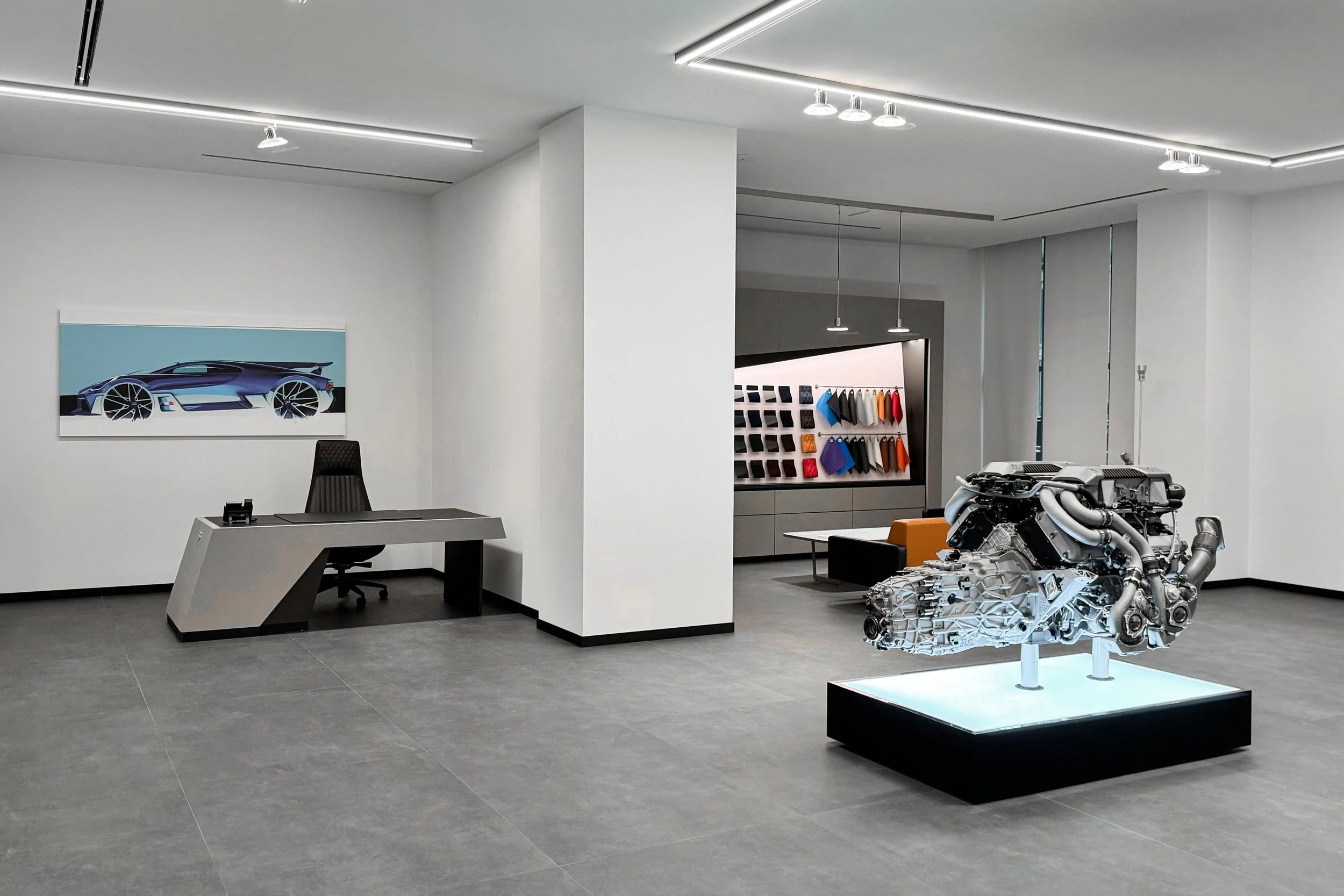 Modern showroom with a car engine on display, an office desk with a chair, and automotive fabric samples on the wall