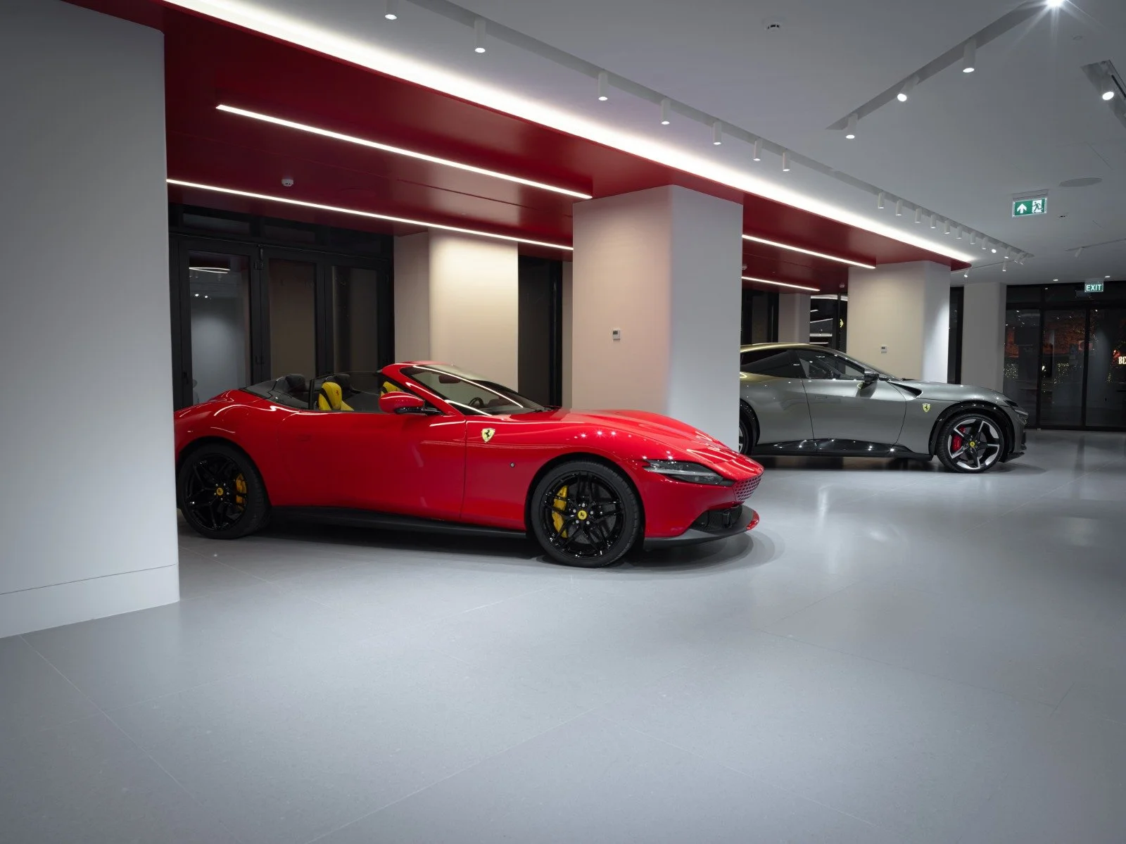 Two luxury sports cars, a red Ferrari convertible and a silver Ferrari coupe, displayed inside a modern showroom with gray flooring and white walls.