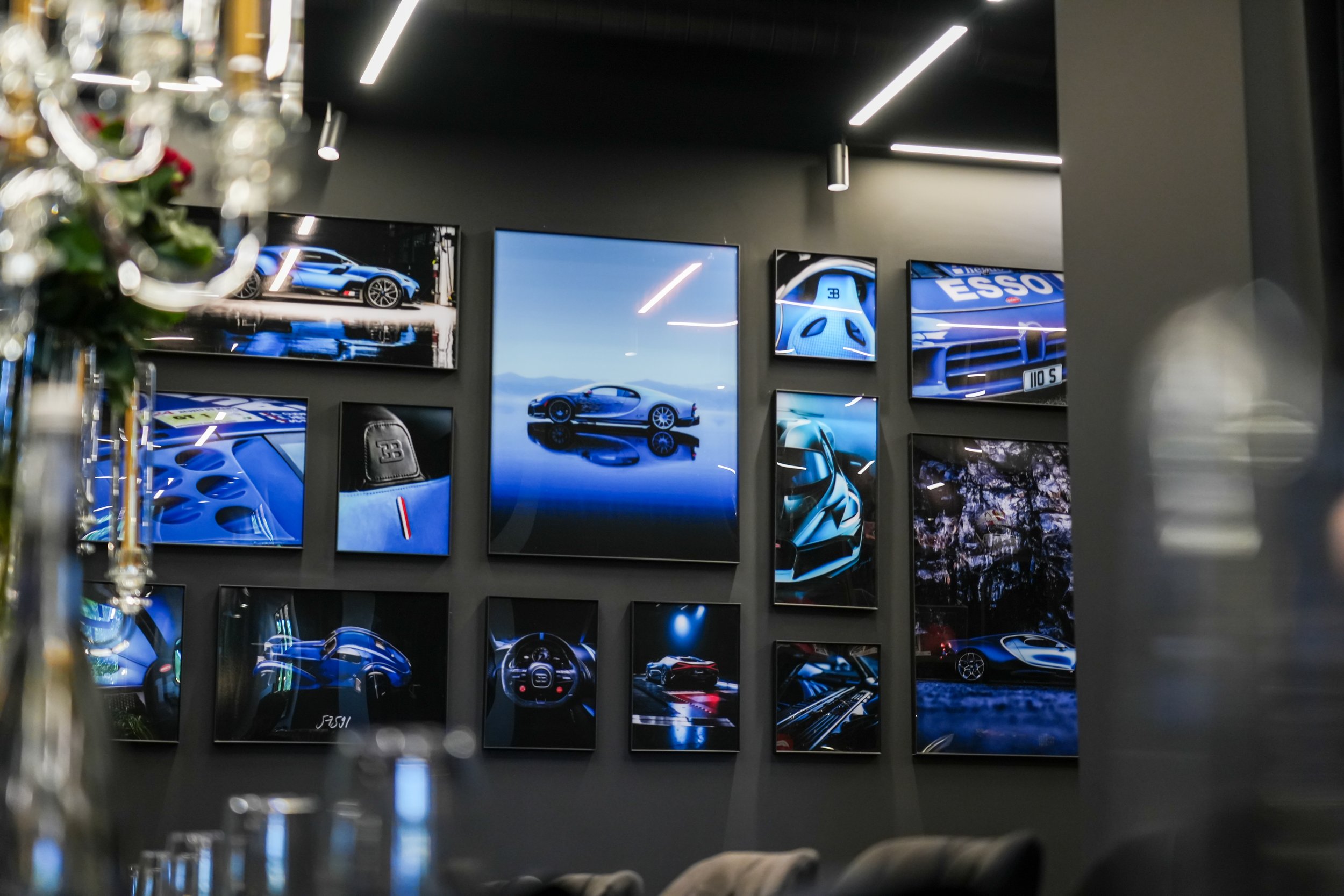 Wall decorated with multiple framed photographs of luxury cars, including a Bugatti, displayed in a modern showroom or gallery. The photos feature various angles and close-ups of sports cars in blue and black.