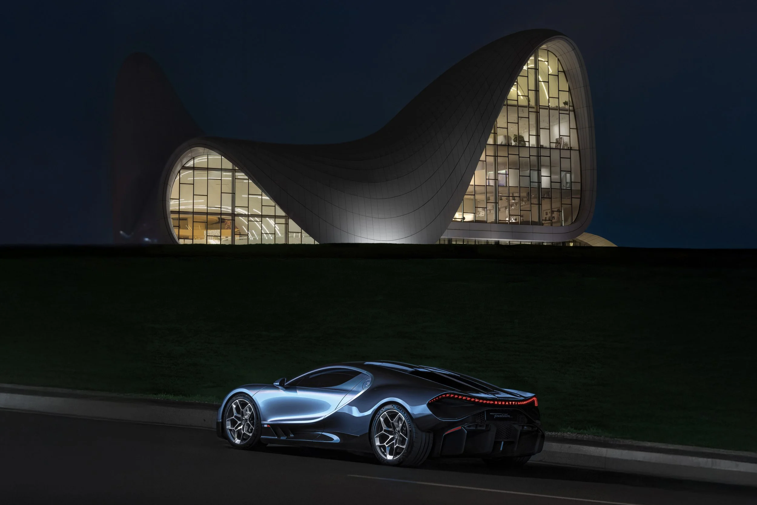 A sleek, modern sports car parked at night in front of a futuristic building with large glass windows and an undulating roof.