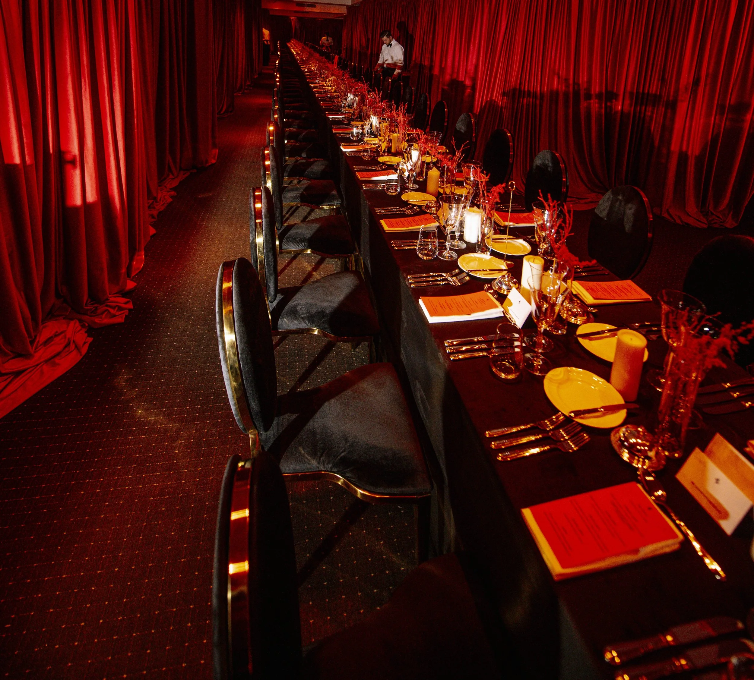 Long banquet table set with plates, glasses, silverware, candles, and menus in an elegant room with red curtains.