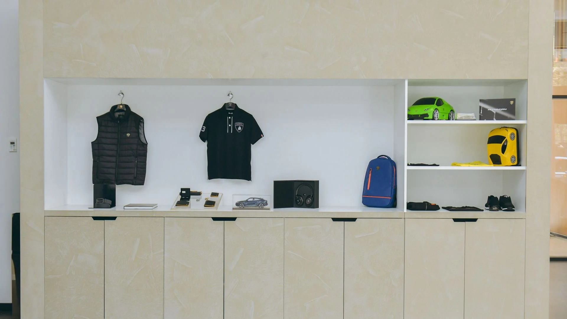 Display shelf in a luxury showroom with clothing, toys, and accessories, including a green toy car, a yellow toy car, a backpack, and small model cars.