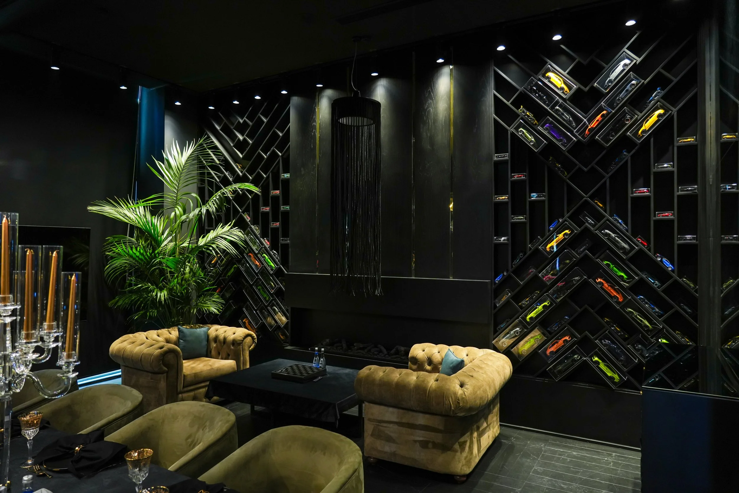 Luxurious lounge with beige velvet sofas, black table, plant, and a wall display of model cars in illuminated cases.