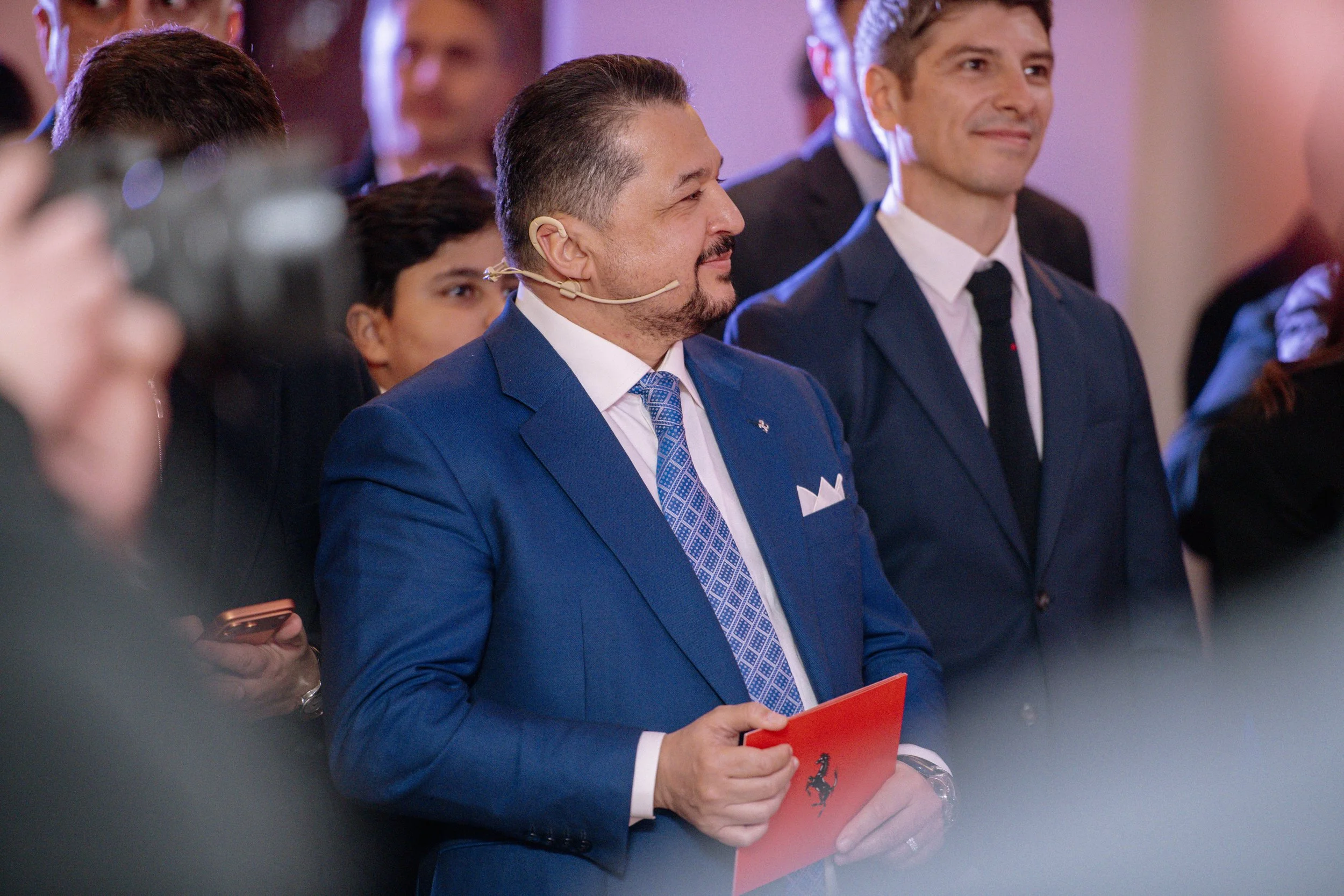A man with a beard and mustache in a blue suit and patterned tie, holding a red folder with a black prancing horse logo, standing among other suited individuals at an indoor event.