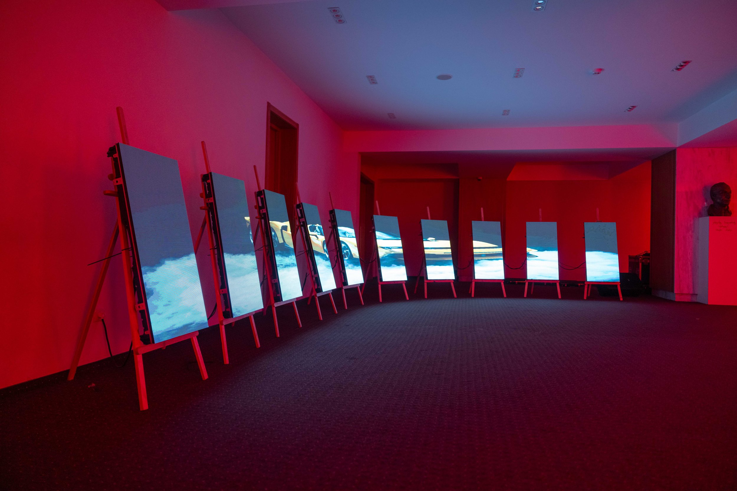 A sleek, blue Bugatti sports car inside a modern showroom with white and black walls, gray flooring, and a large black display screen with the white text 'BUGATTI'.