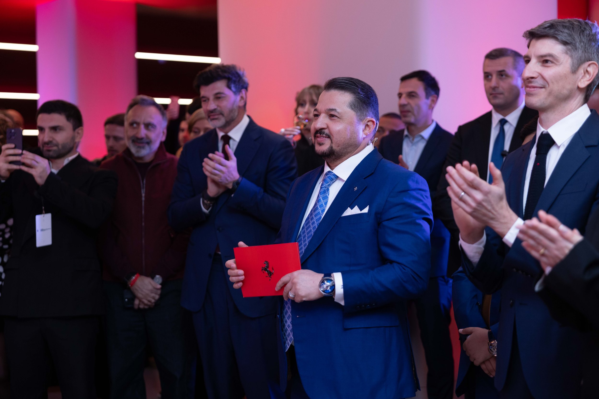 A formal dining table set for an event, decorated with lit candles, floral arrangements, wine glasses, and tableware, with people seated in the background at a dark indoor venue.