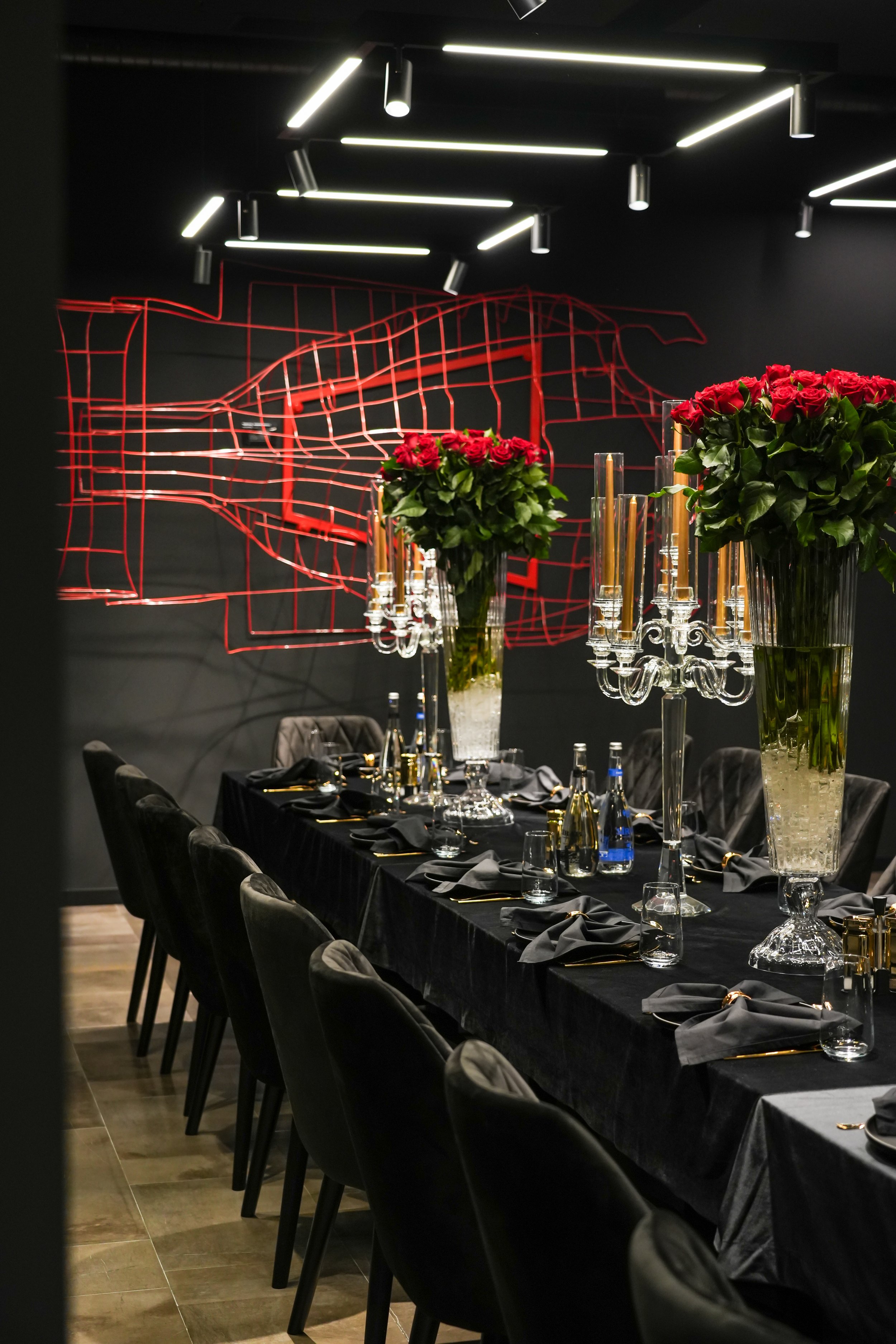 Elegant dining room with a long black table set with black napkins, gold flatware, and glassware. Tall vases with pink roses and crystal candelabras are centered on the table. The background features a black wall with an abstract red wire art piece a