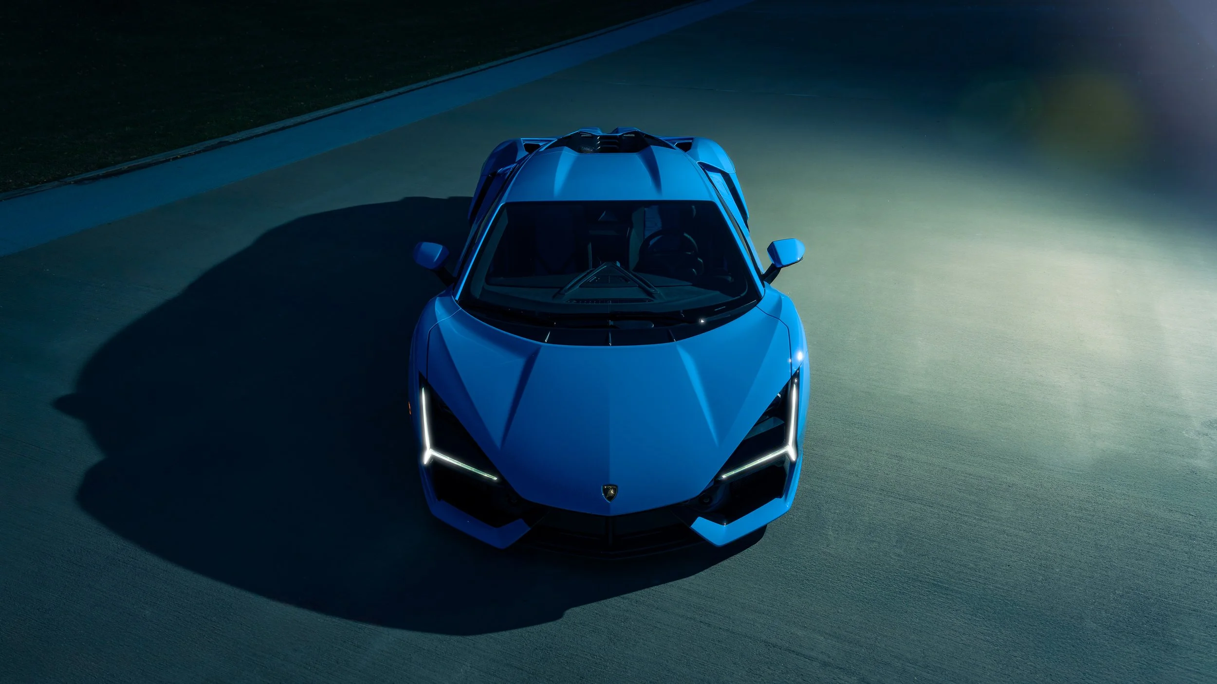 Blue sports car on a racetrack at night, with illuminated front lights and a shadow cast onto the track.