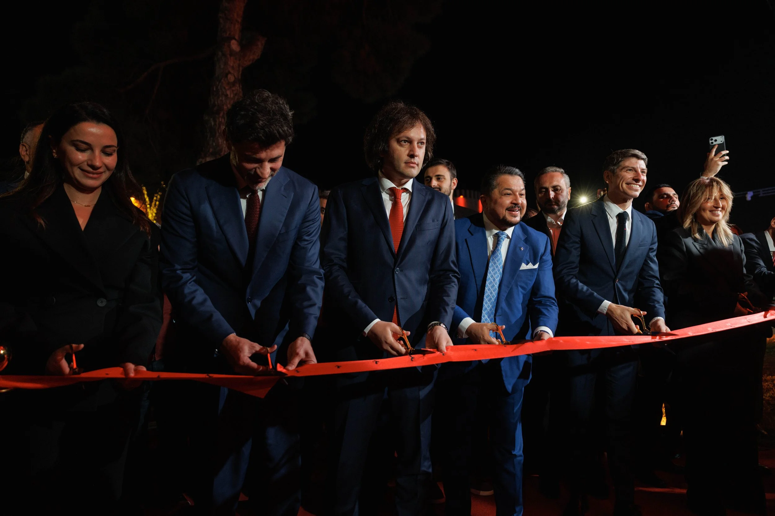 A sleek blue Bugatti sports car with butterfly doors open, parked on a light-colored tiled surface in front of a modern, curved metallic building.