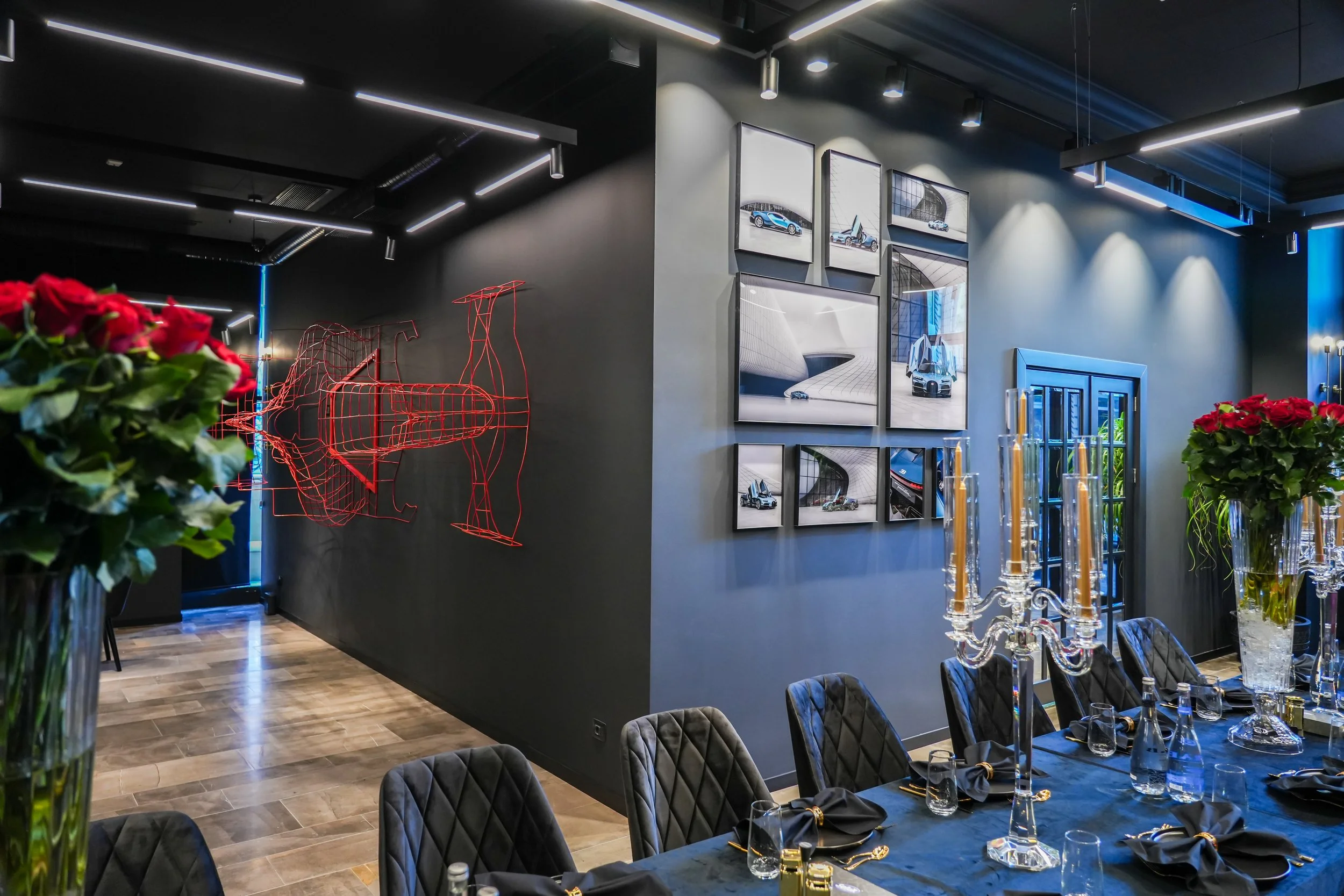 A modern interior room with black walls, featuring a red wireframe sculpture of a racing car on one wall and framed images of cars and architecture on another. There is a long dining table with black chairs, gold and crystal candlesticks, and floral 