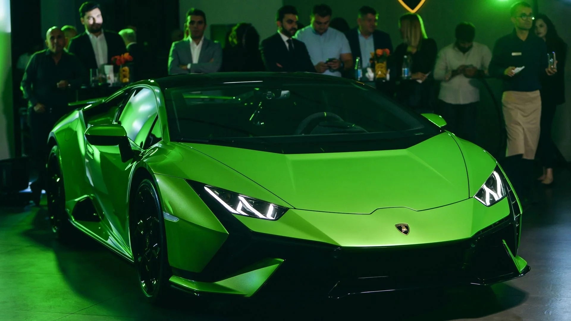 A vibrant green Lamborghini sports car is showcased indoors at a car event, with a group of people observing it from behind, some using their phones.