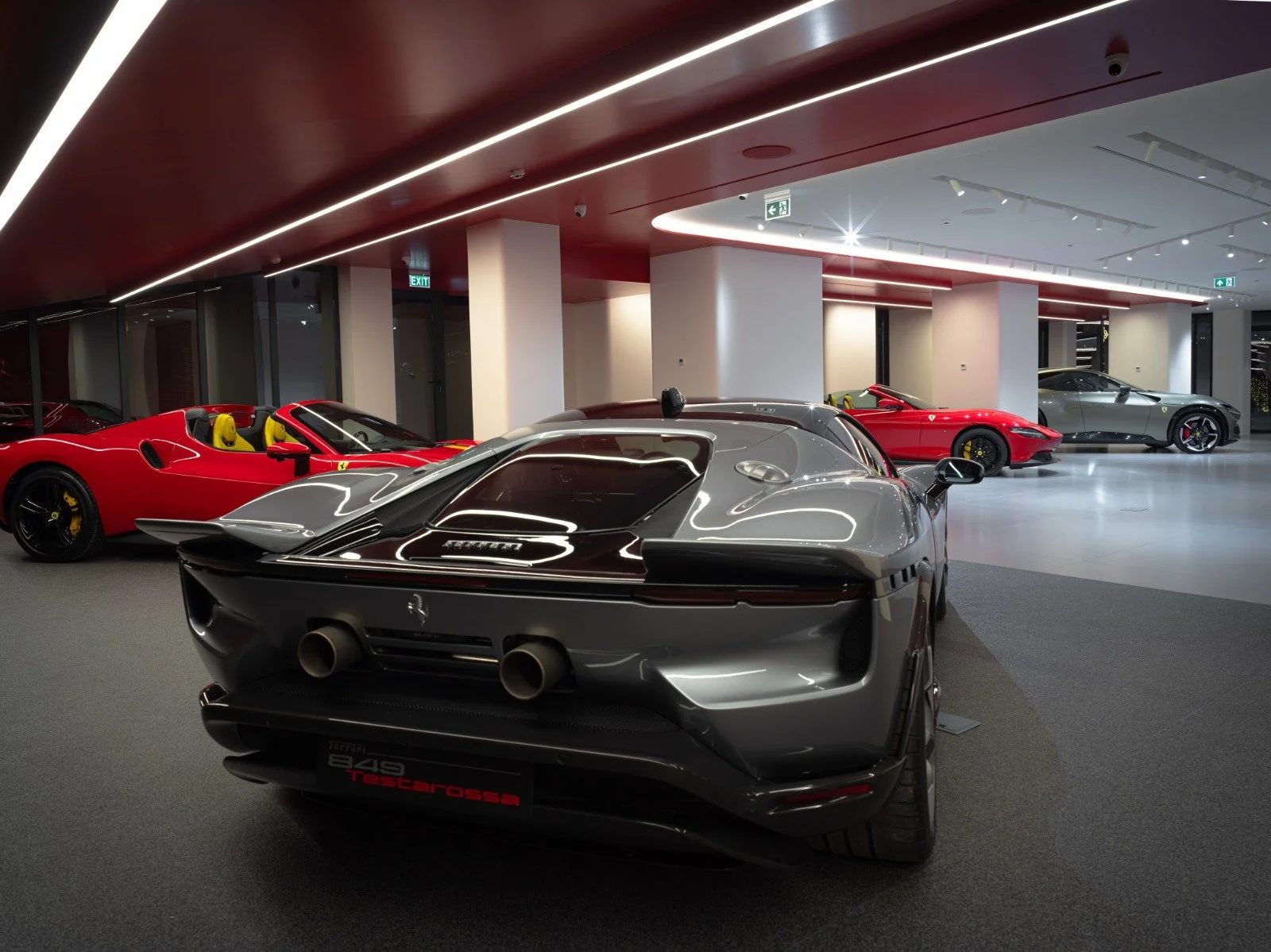 A showroom displaying five Ferrari sports cars, including a silver Ferrari 488 Pista, four red Ferrari convertibles, and a grey Ferrari SUV, in a modern, well-lit space with a red ceiling and white walls.