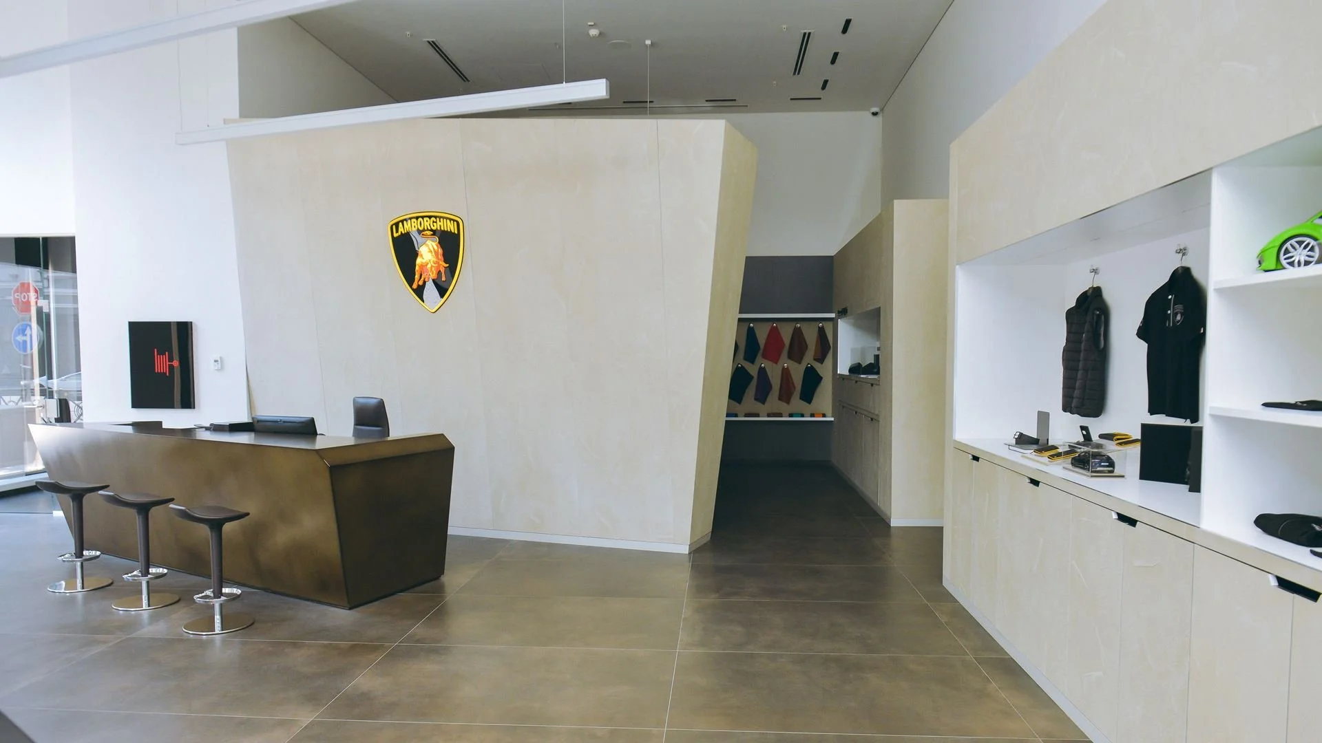 Modern Lamborghini showroom reception area with a sleek dark brown reception desk, three black bar stools, Lamborghini logo on the wall, and display shelves with jackets and car accessories.