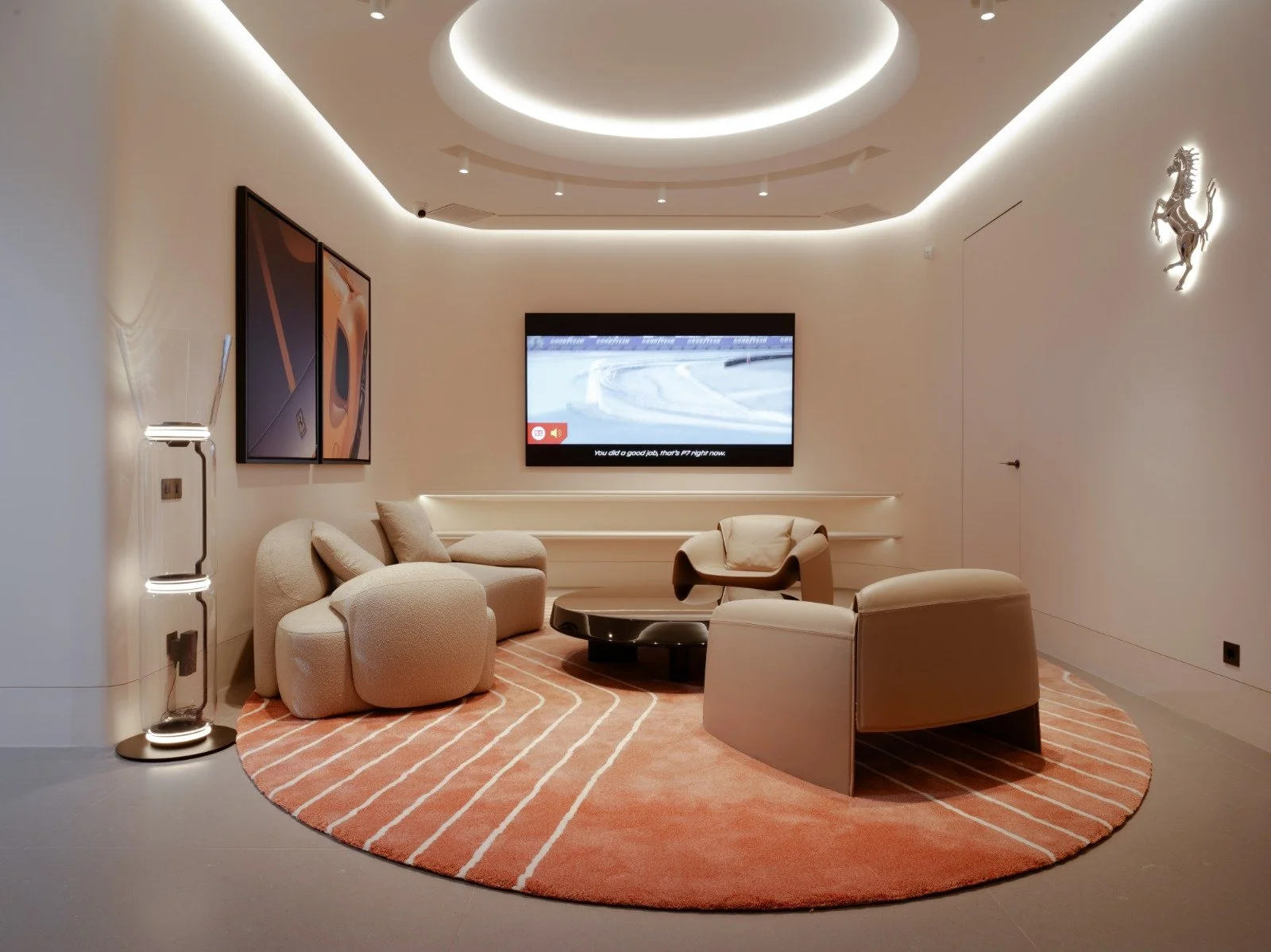 Modern hotel lounge with beige seating, a round pink rug with white lines, a black coffee table, wall-mounted TV showing a race car track, two sleek wall art pieces, a Ferrari logo on the wall, and modern lighting fixtures.