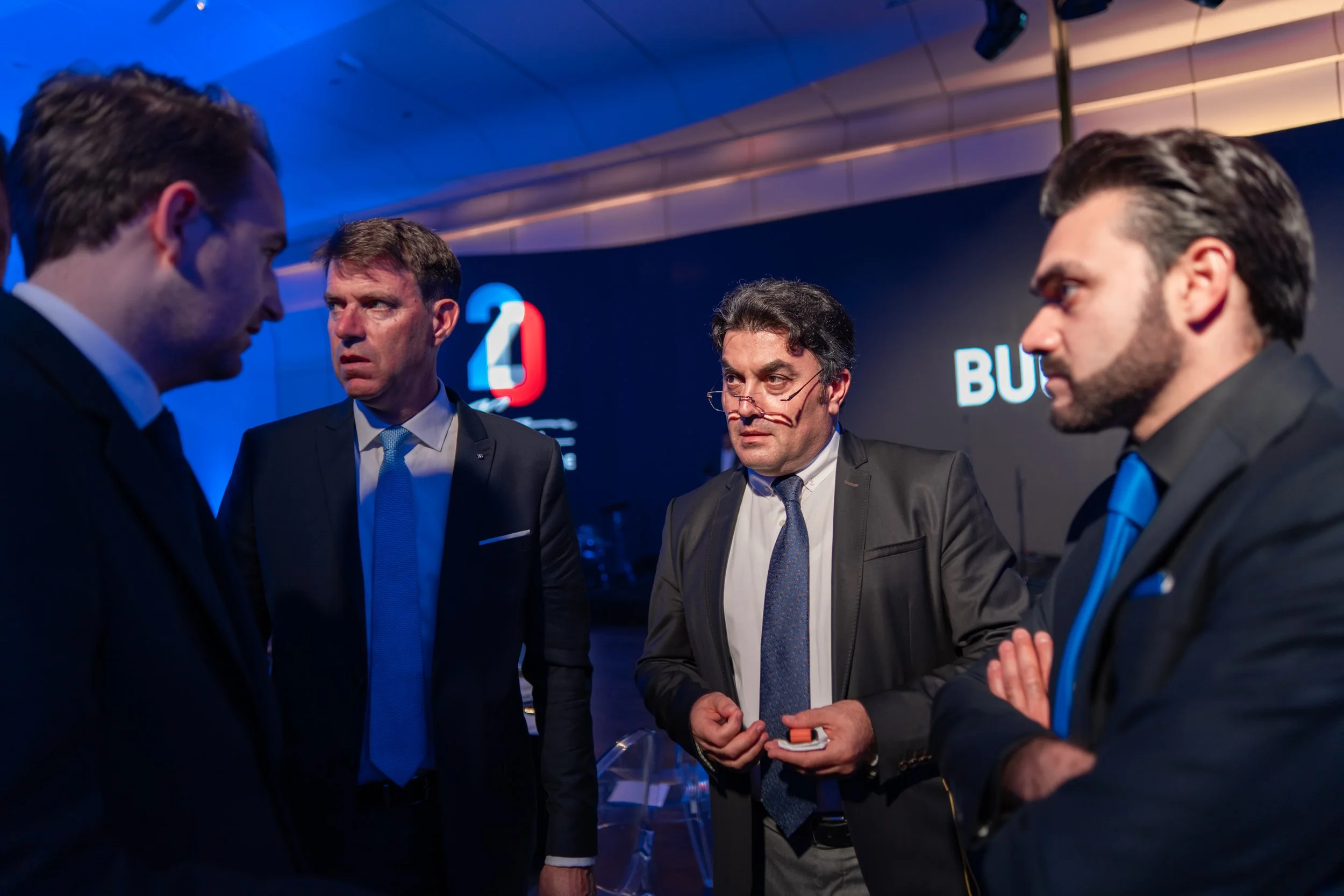 Four men in suits having a serious conversation at a formal event, with illuminated signs and a blue backdrop in the background.