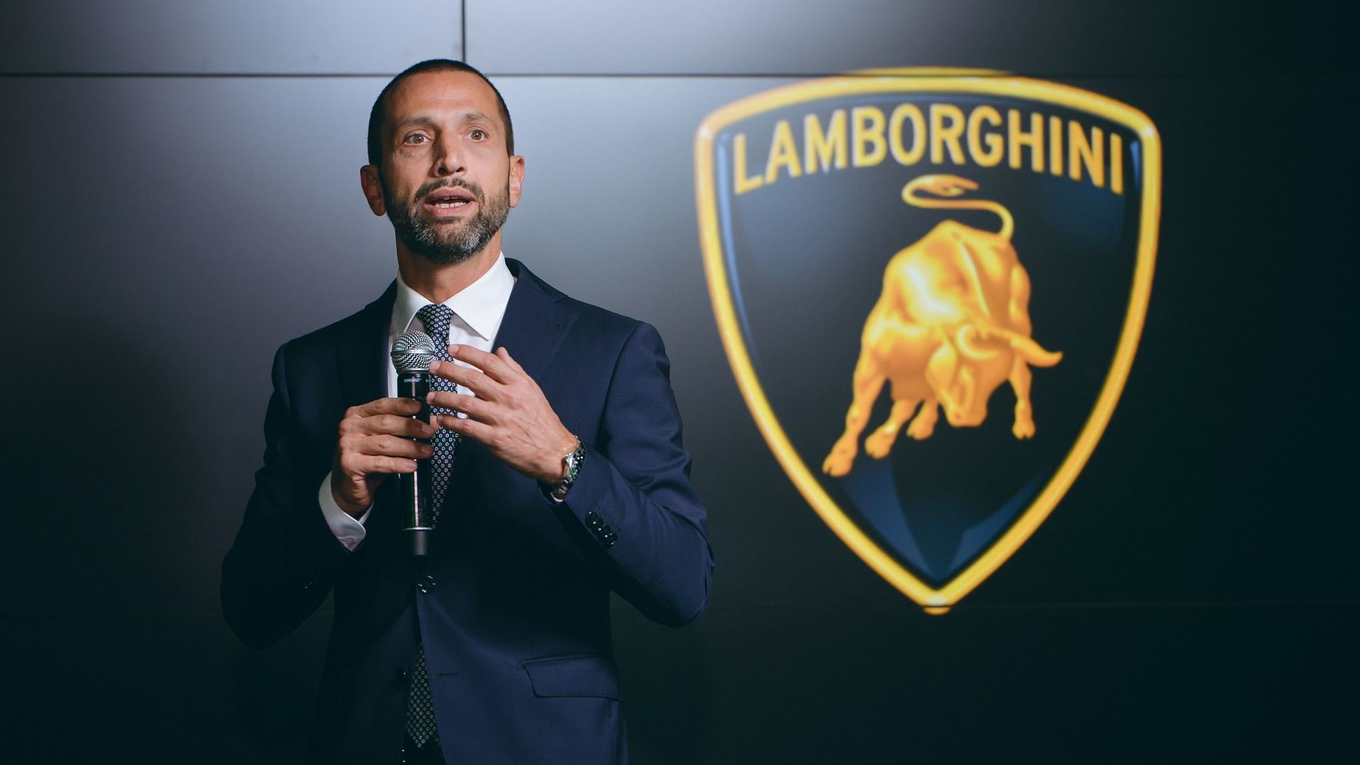 Man in a navy suit giving a speech, holding a microphone, with Lamborghini logo in the background.